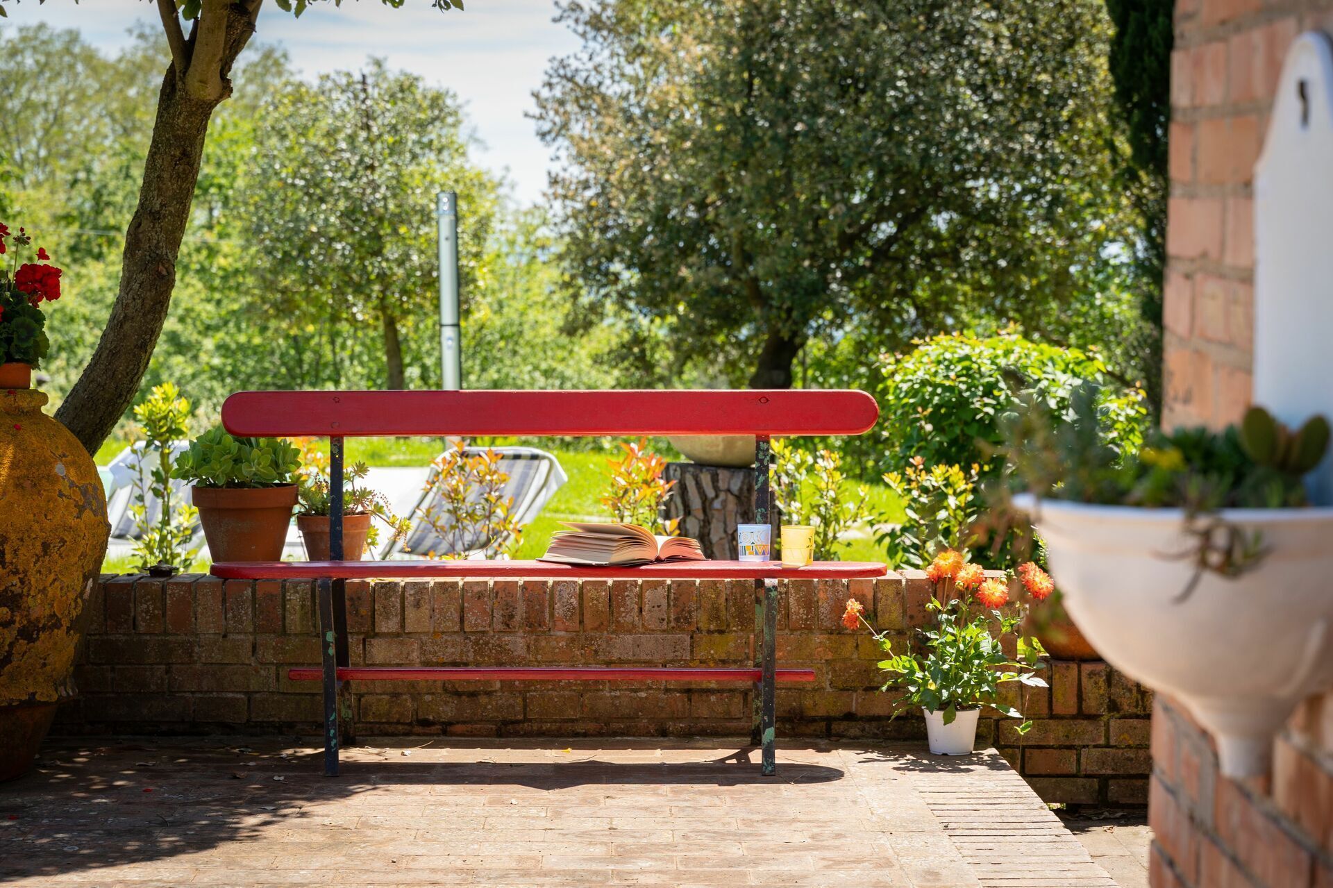 Un banco rojo con un libro y una taza, rodeado de macetas con plantas y un muro de ladrillo.