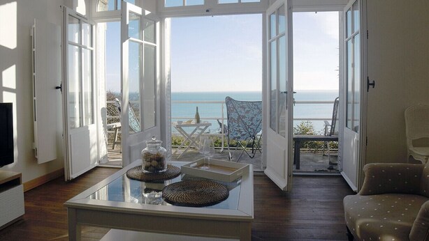 Une pièce spacieuse avec de grandes fenêtres donnant sur la mer. On y trouve une table en verre agrémentée d’objets décoratifs et une chaise.