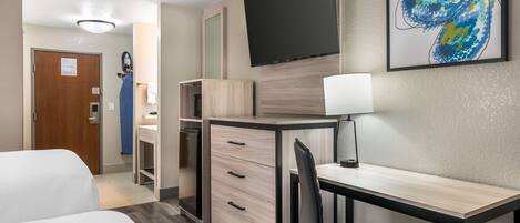 A hotel room with a bed, a desk, a chair, a TV, and a painting on the wall.