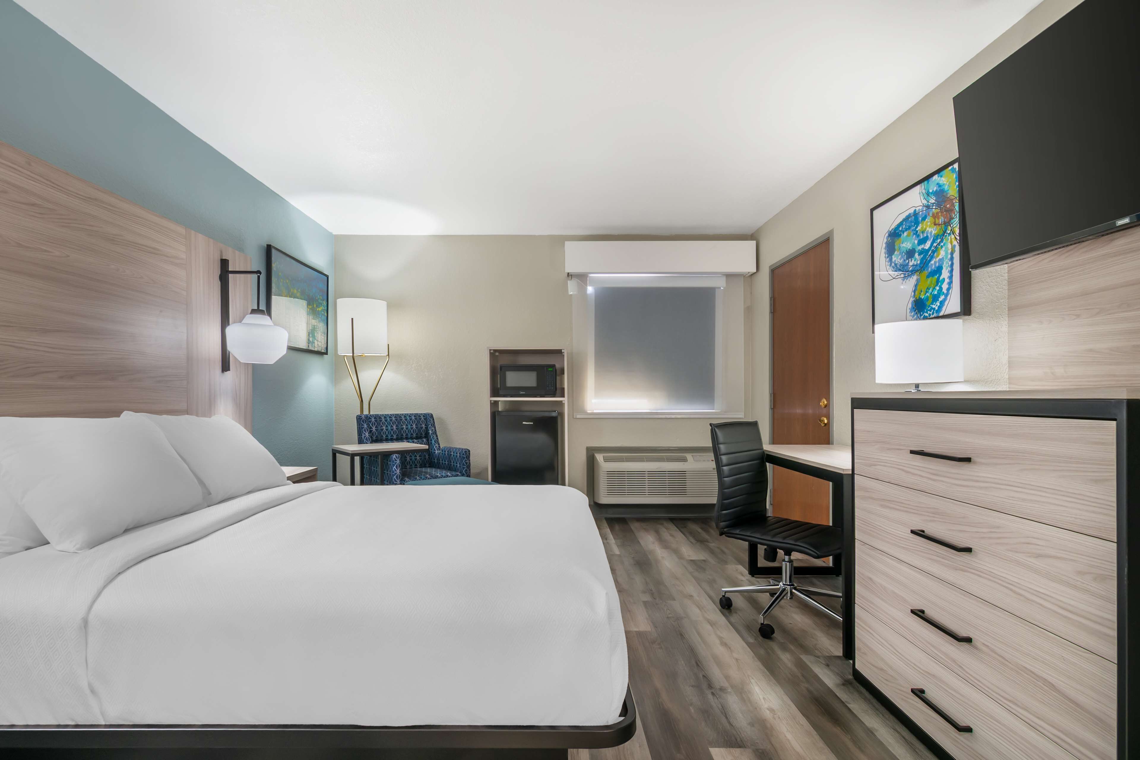 A hotel room with a bed, desk, chair, dresser, and artwork.