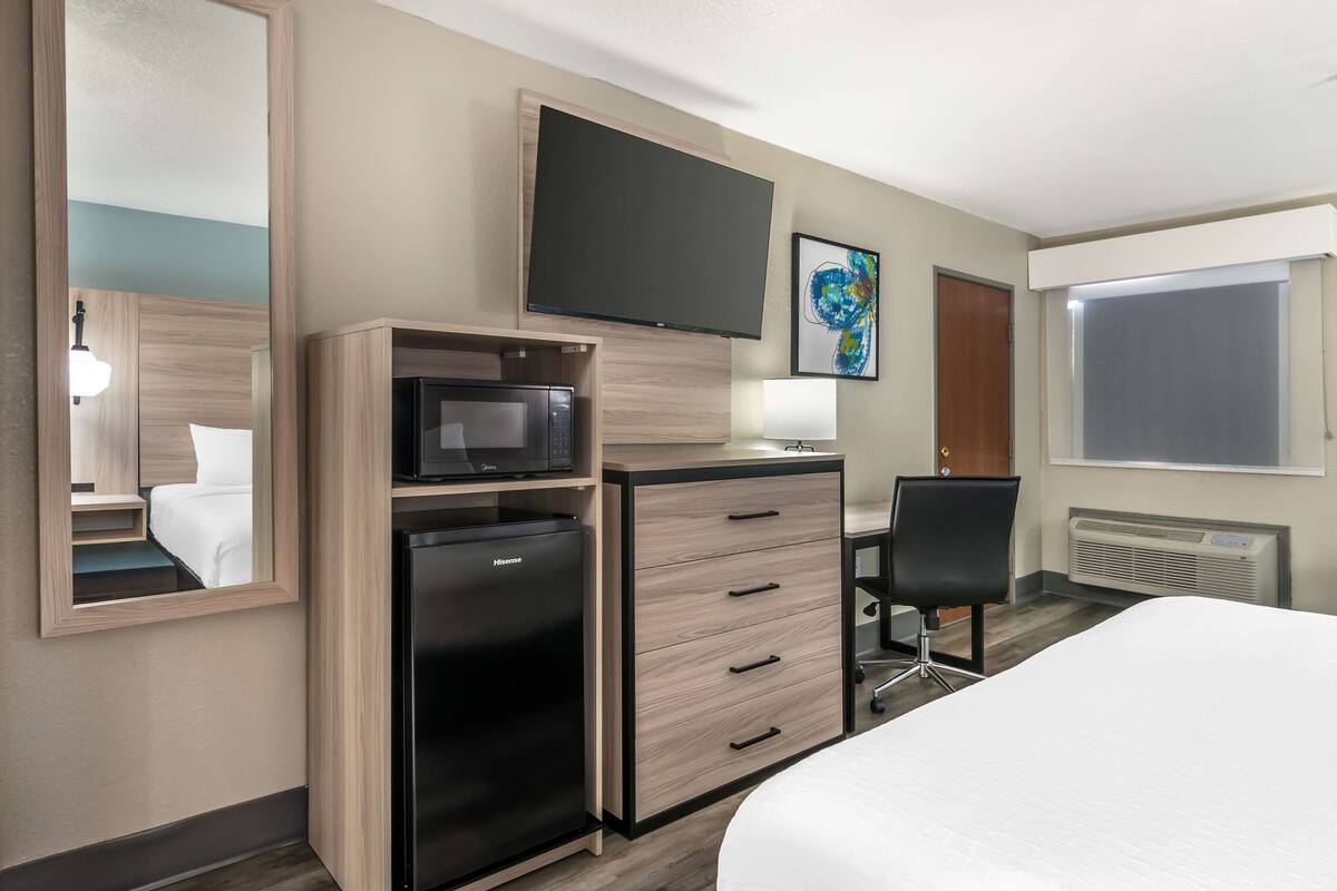 A hotel room with a bed, a desk with a computer, a refrigerator, a microwave, a flat-screen TV, a chair, and a window.
