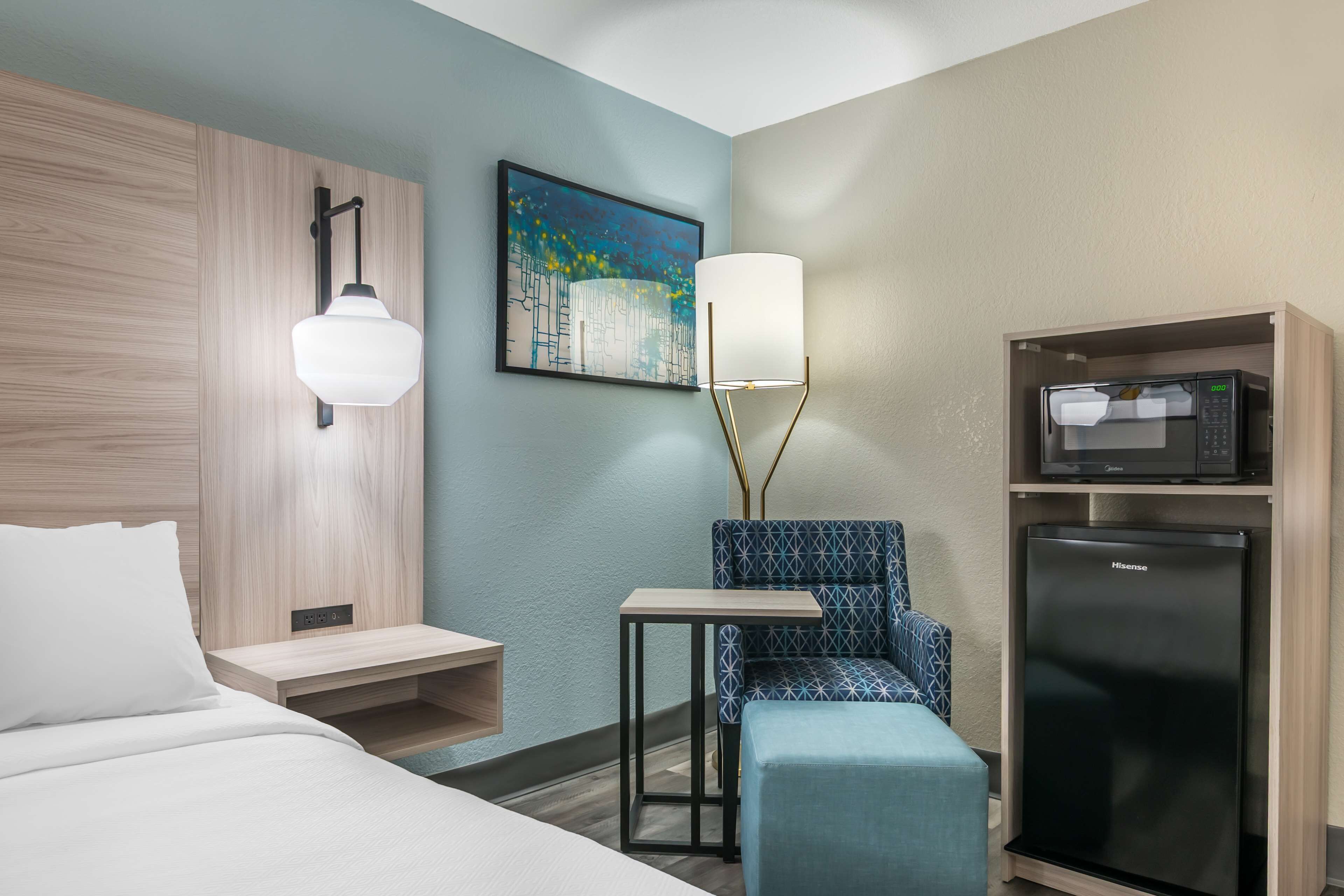A hotel room with a bed, a desk, a chair, a microwave, and a refrigerator.