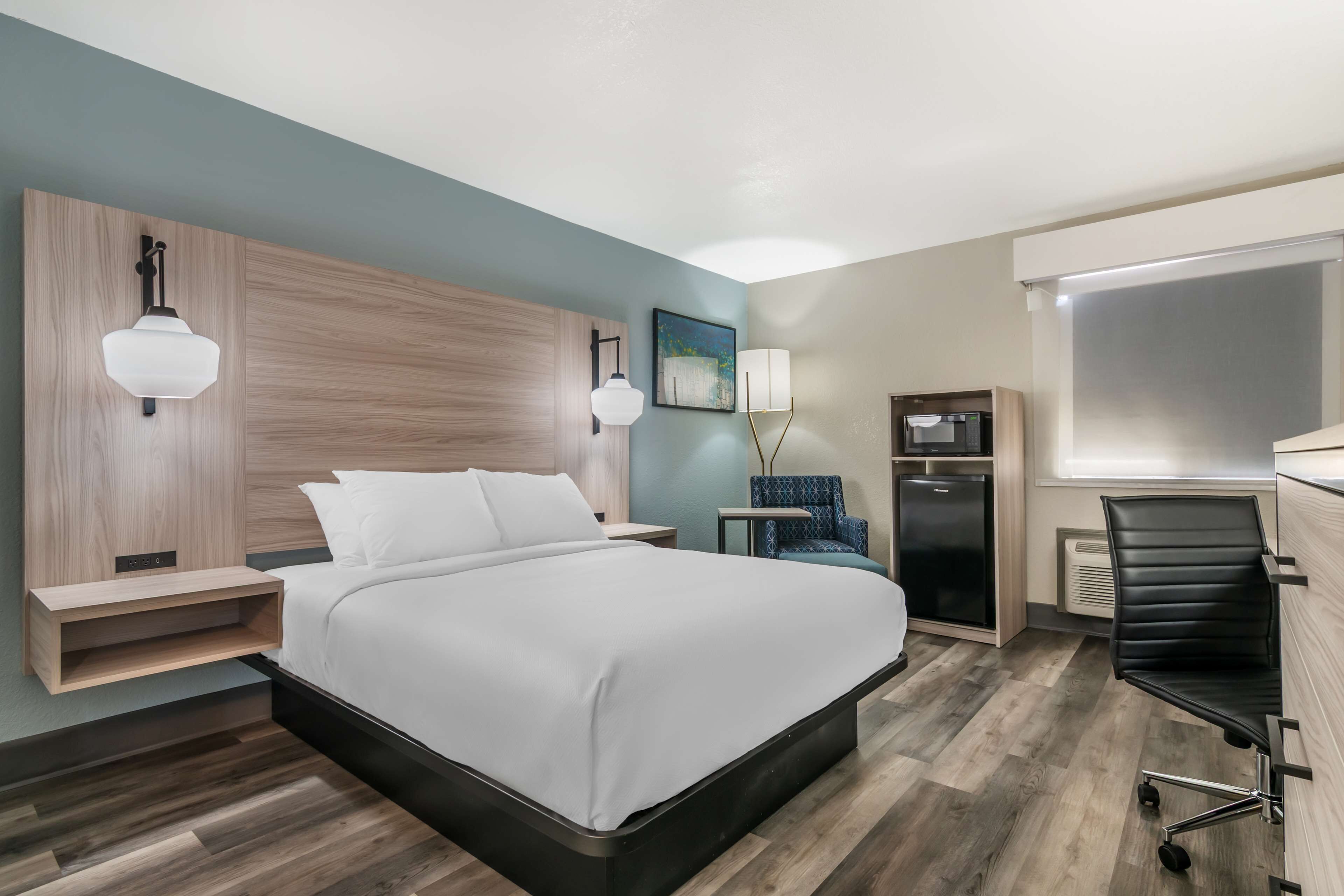 Una habitación de hotel moderna con una cama grande, mesitas de noche, un escritorio, una silla y un refrigerador.
