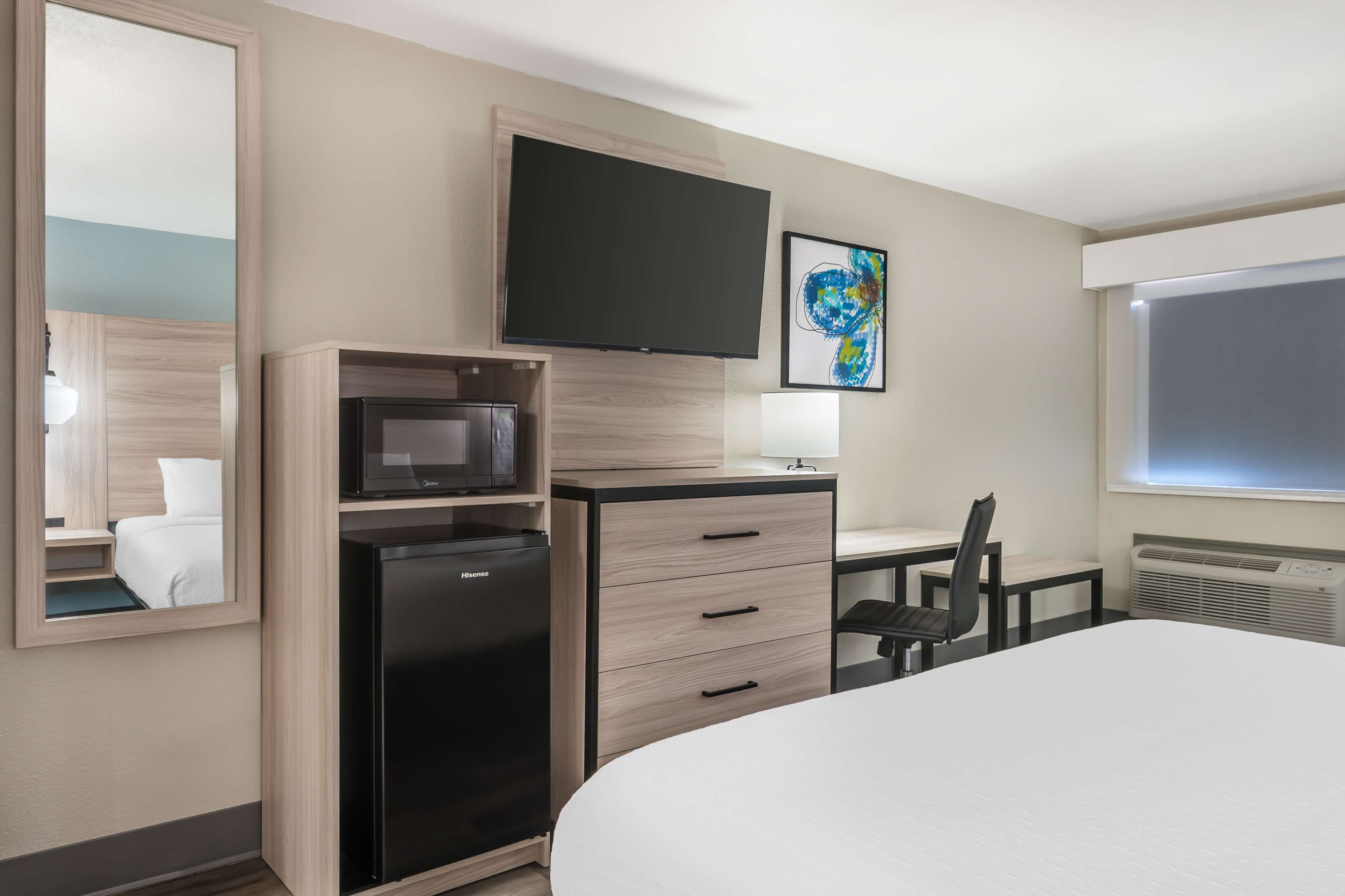 A hotel room with a bed, a desk with a chair, a flat-screen TV, a microwave, a refrigerator, a large mirror, and a framed picture on the wall.