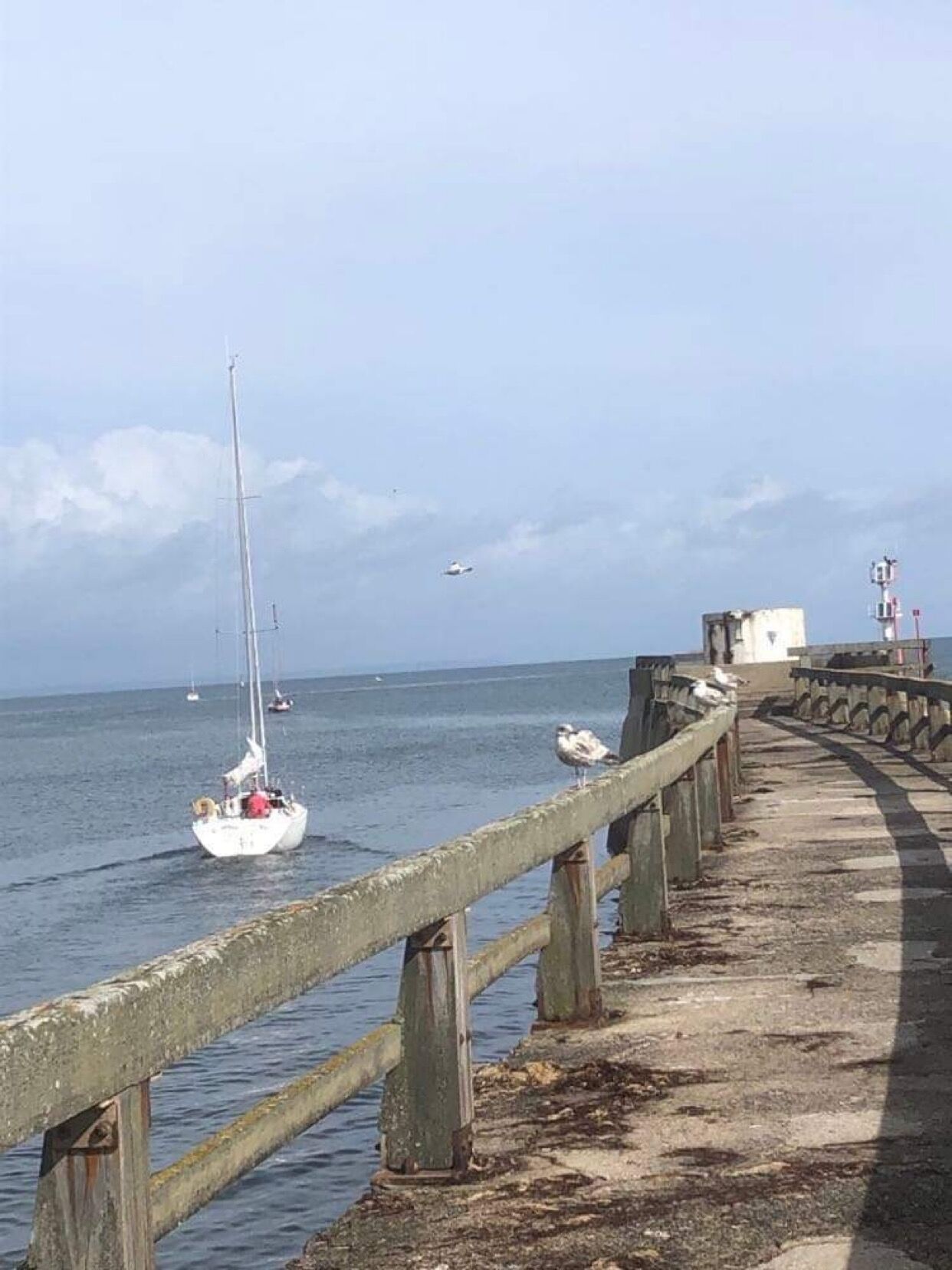 Une jetée avec une rambarde en bois, un voilier sur l’eau et un phare au loin.
