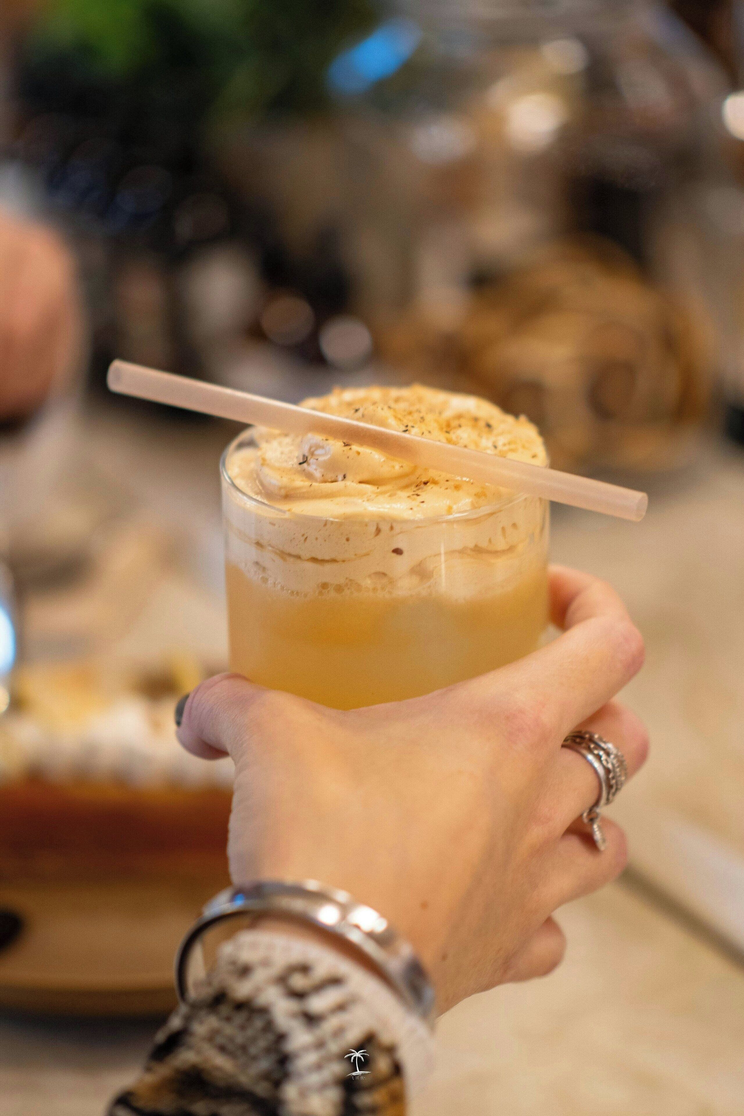 Una persona tiene un bicchiere con una bevanda a strati, guarnita con panna montata e una cannuccia.