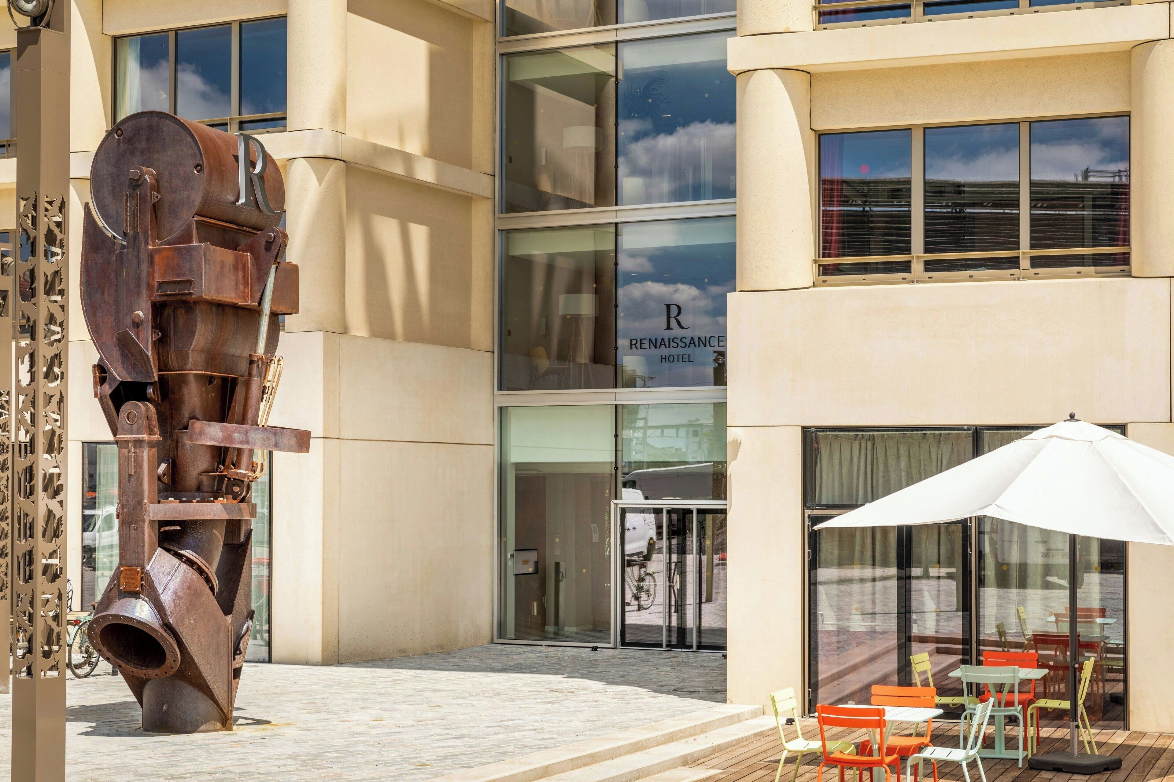 A modern building with a large metal sculpture in front, a glass door entrance, and outdoor seating with umbrellas.