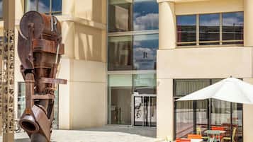 Un bâtiment moderne, avec une grande sculpture métallique à l’avant, une entrée avec une porte en verre et un espace extérieur aménagé avec des parasols.