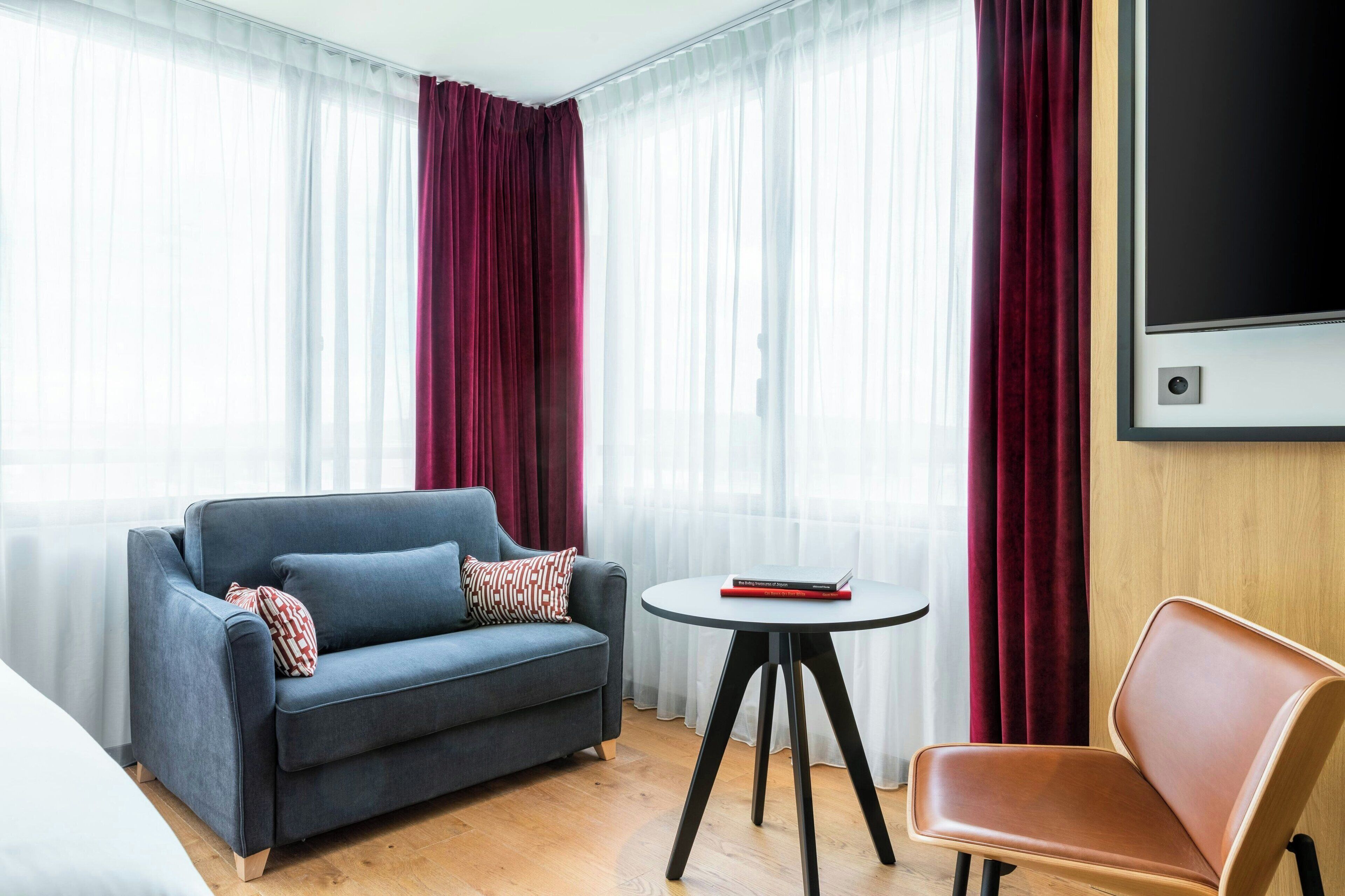A modern hotel room with a grey sofa, a small round table, a brown chair, and a large TV screen.