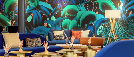 Un espacio lounge moderno con un conjunto de sofá azul, mesas redondas doradas y un vibrante mural tropical de fondo.