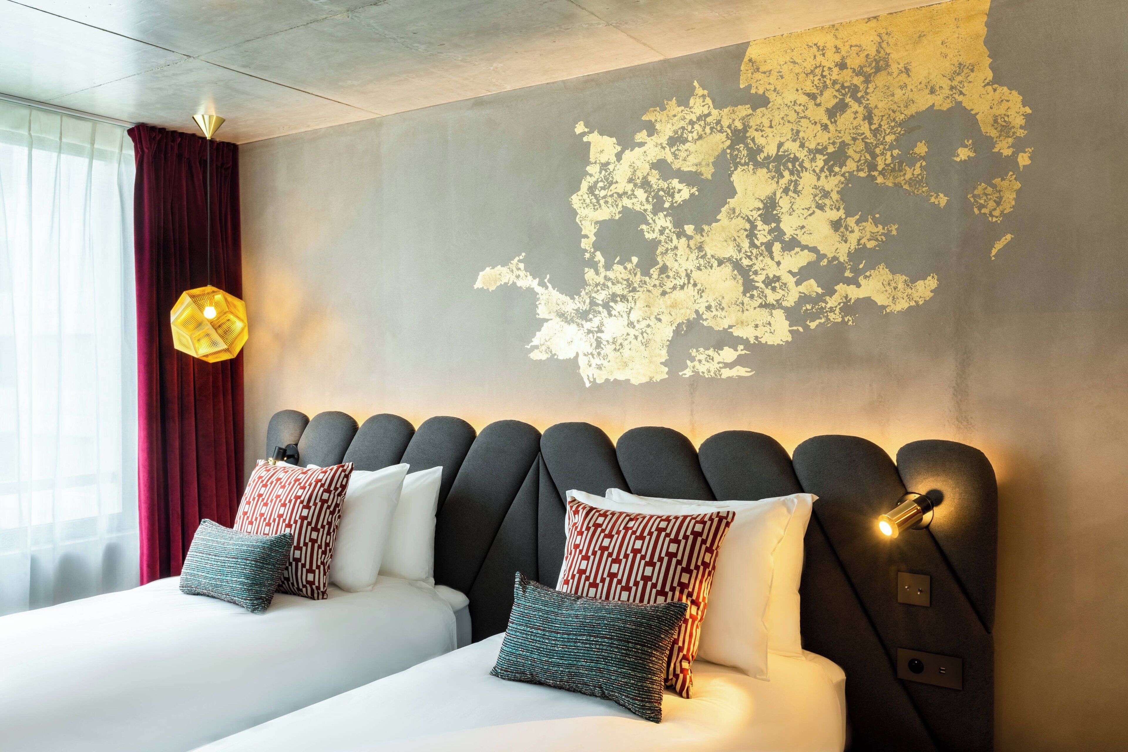 A hotel room with a modern headboard, two beds, patterned pillows, a large window with curtains, and a textured wall.