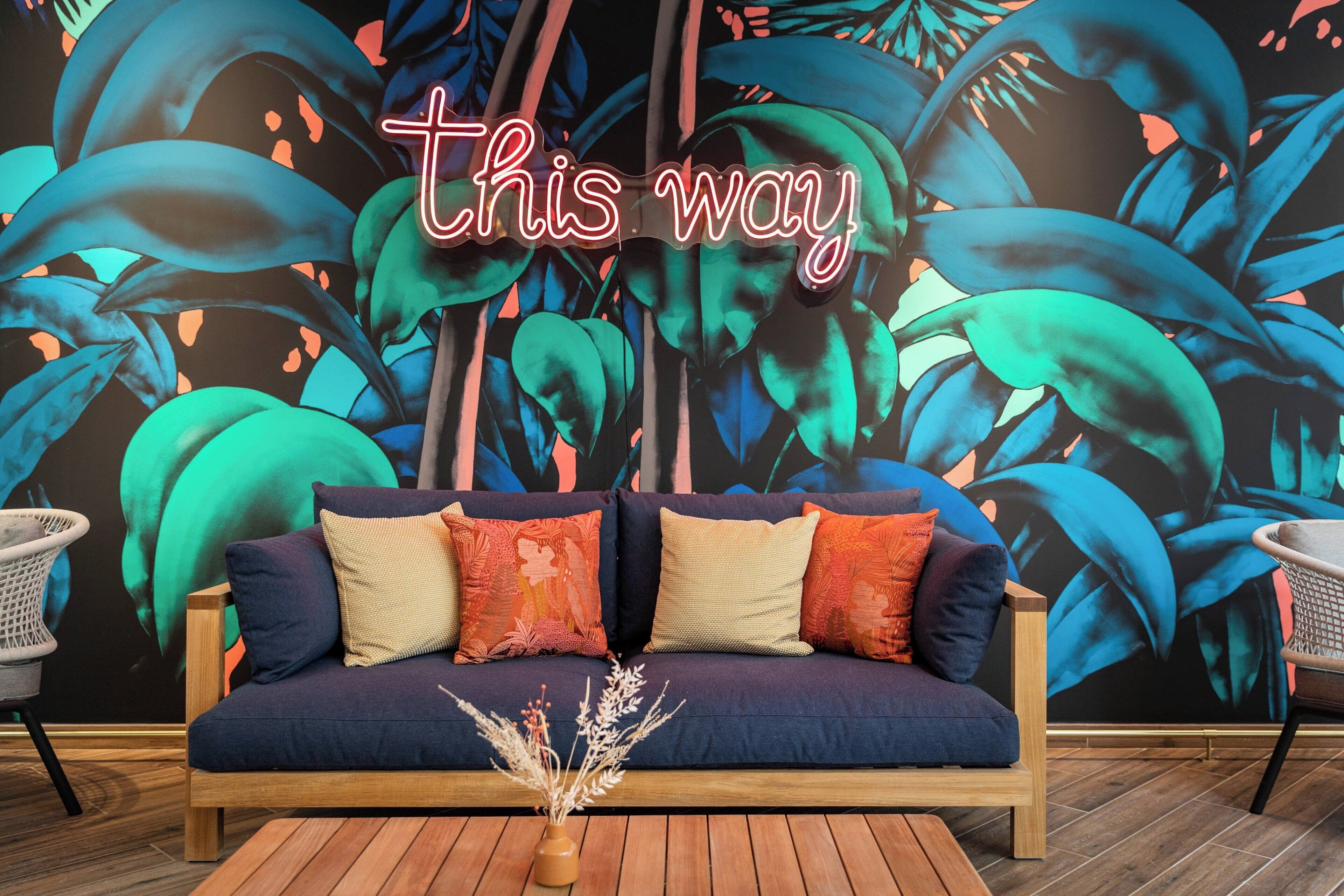 Een moderne woonkamer met een donkerblauwe bank, meerdere kussens en een neonbord met de tekst 'this way' voor een levendig tropisch behang.