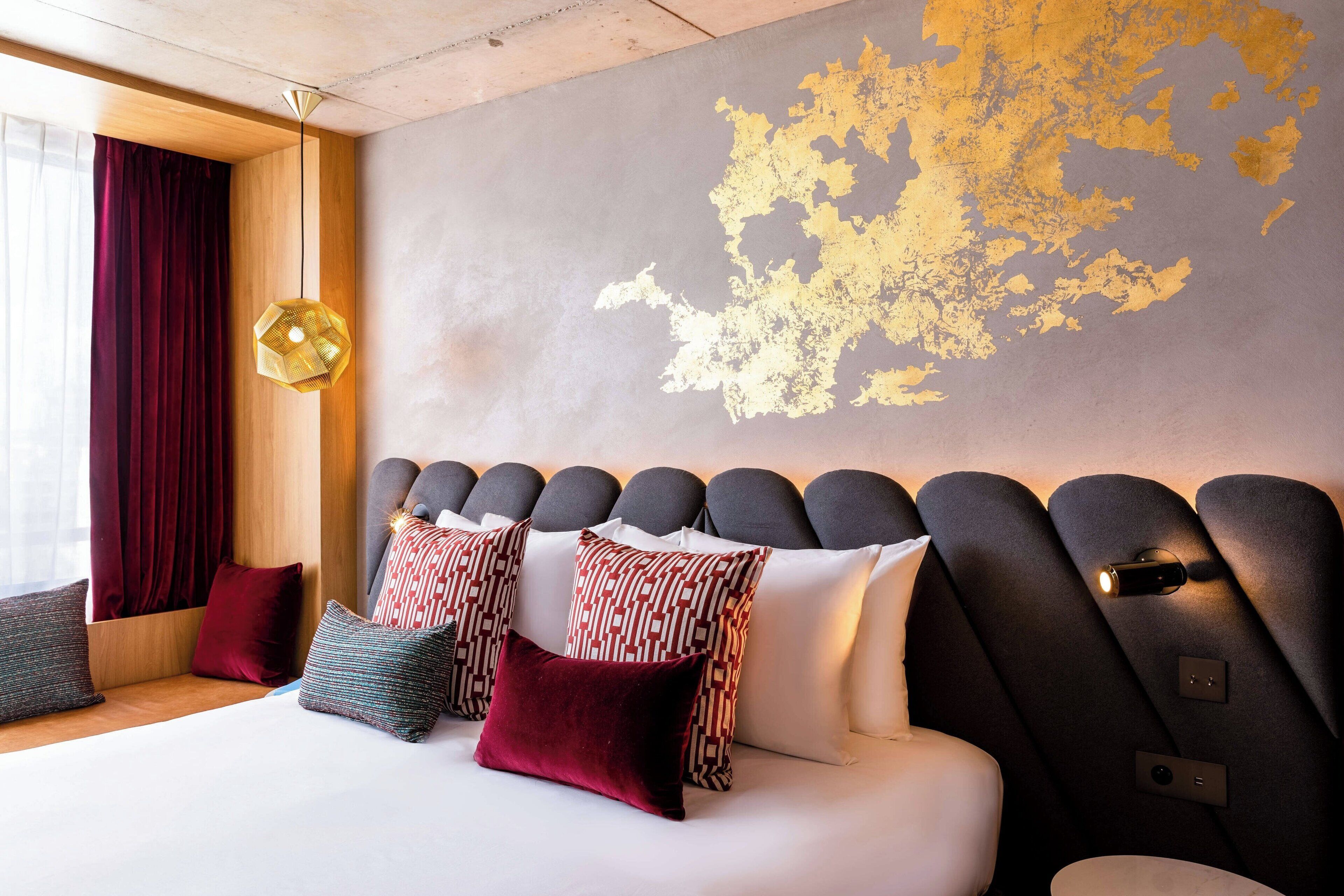 A modern hotel room with a large bed, patterned pillows, a unique wall design, and a pendant light.