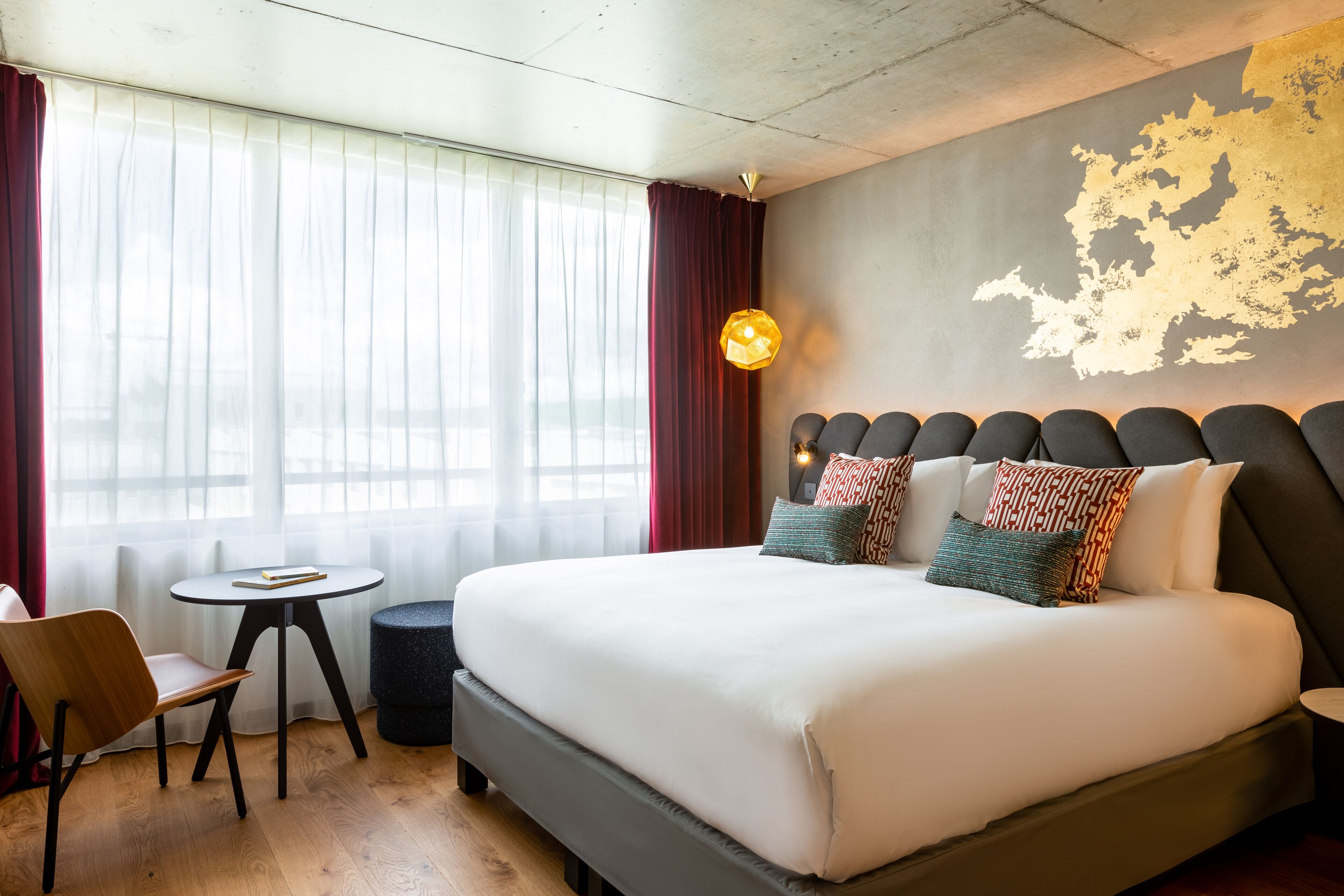 Ein Hotelzimmer mit einem großen Bett, einem kleinen runden Tisch, einem Stuhl und Blick auf die Stadt durch große Fenster.