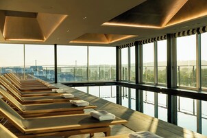 Un espacio interior iluminado por el sol, con filas de tumbonas de madera y amplias ventanas que dan a un puente y al agua.