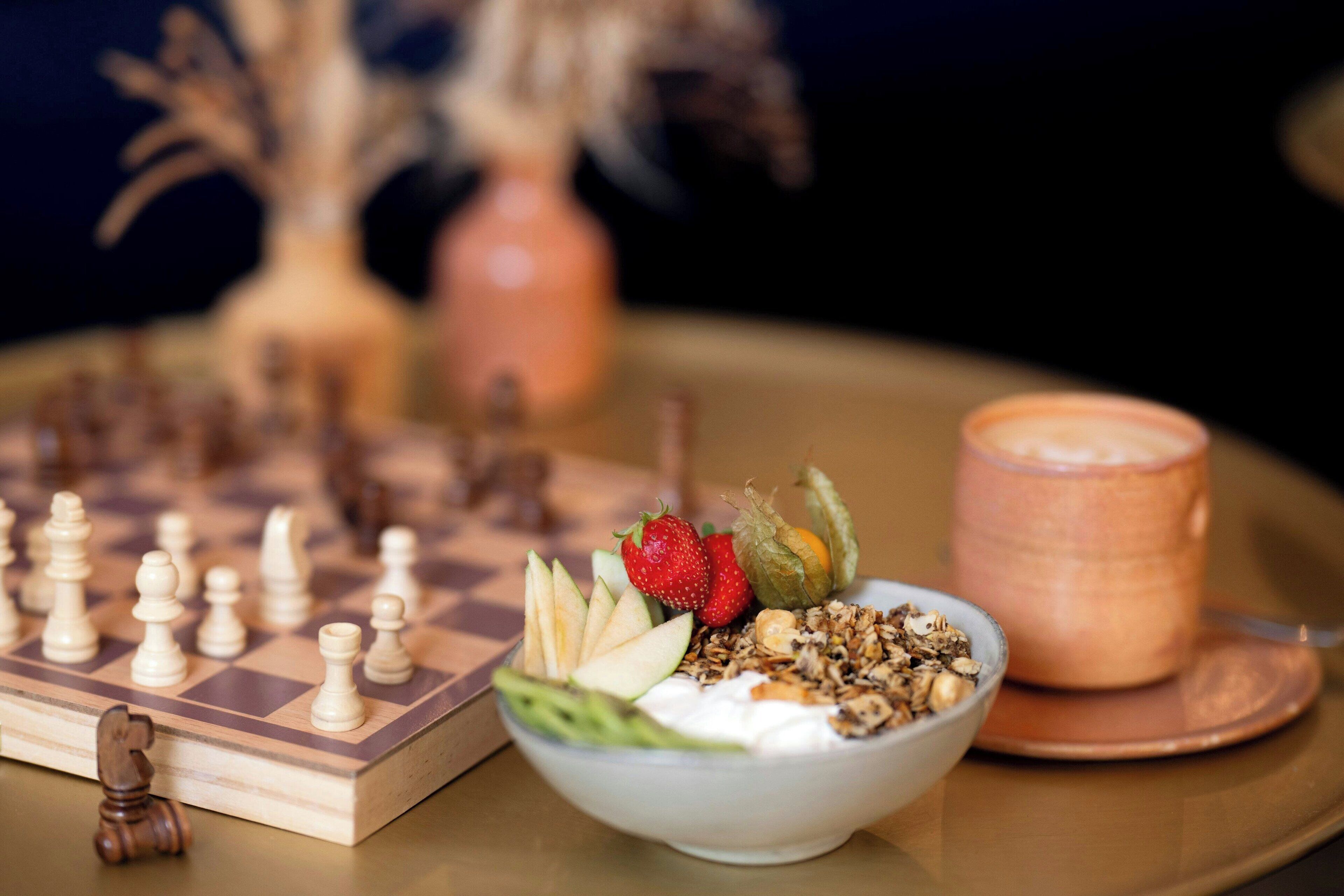 A chessboard, a bowl of yogurt with fruits and granola, and a cup of coffee on a table.