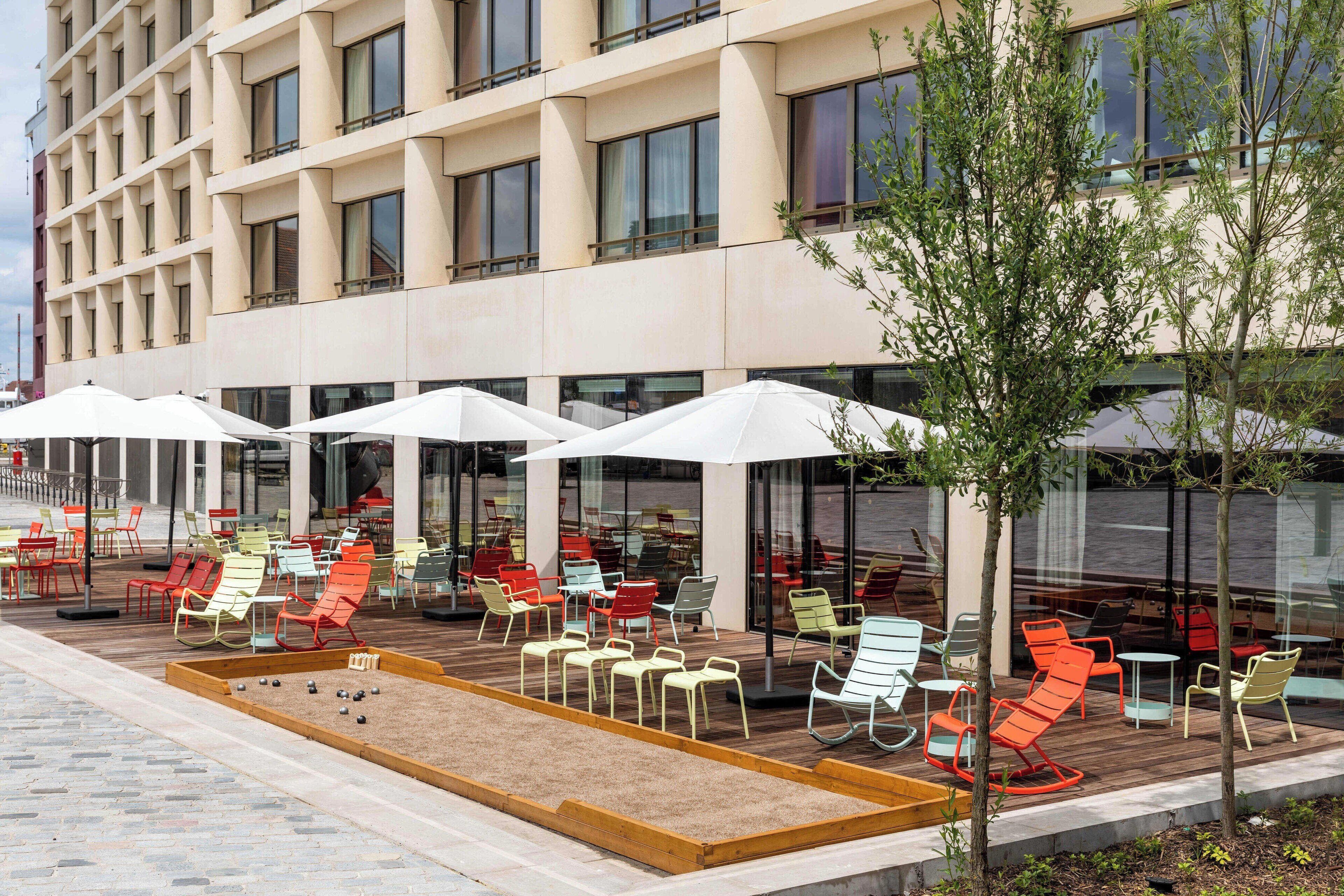 An outdoor seating area with colorful chairs and umbrellas, a sandbox, and a building with large windows in the background.