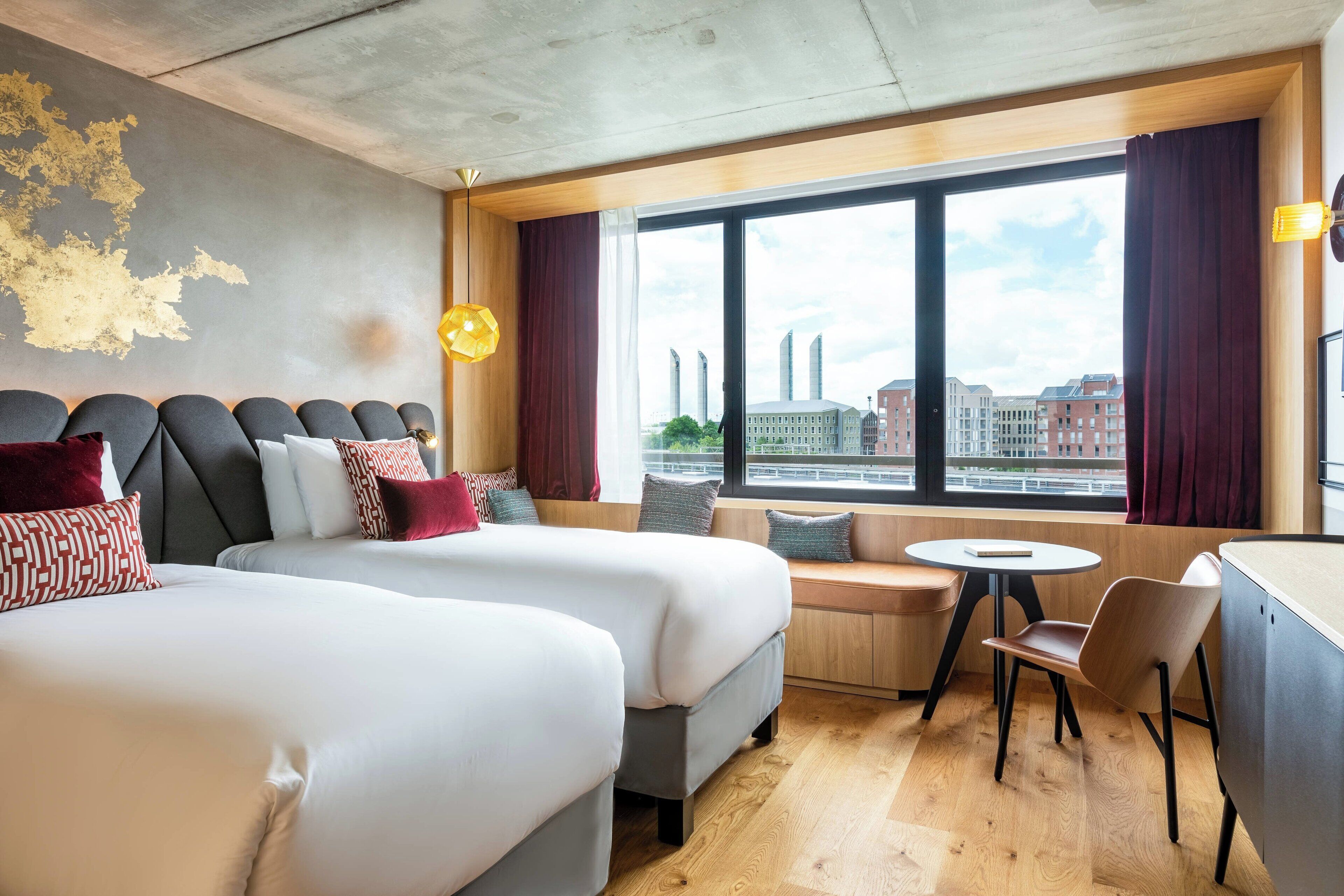 Habitación de hotel con dos camas, una mesita y una silla. Se aprecia un ventanal con vistas a edificios y al paisaje urbano.