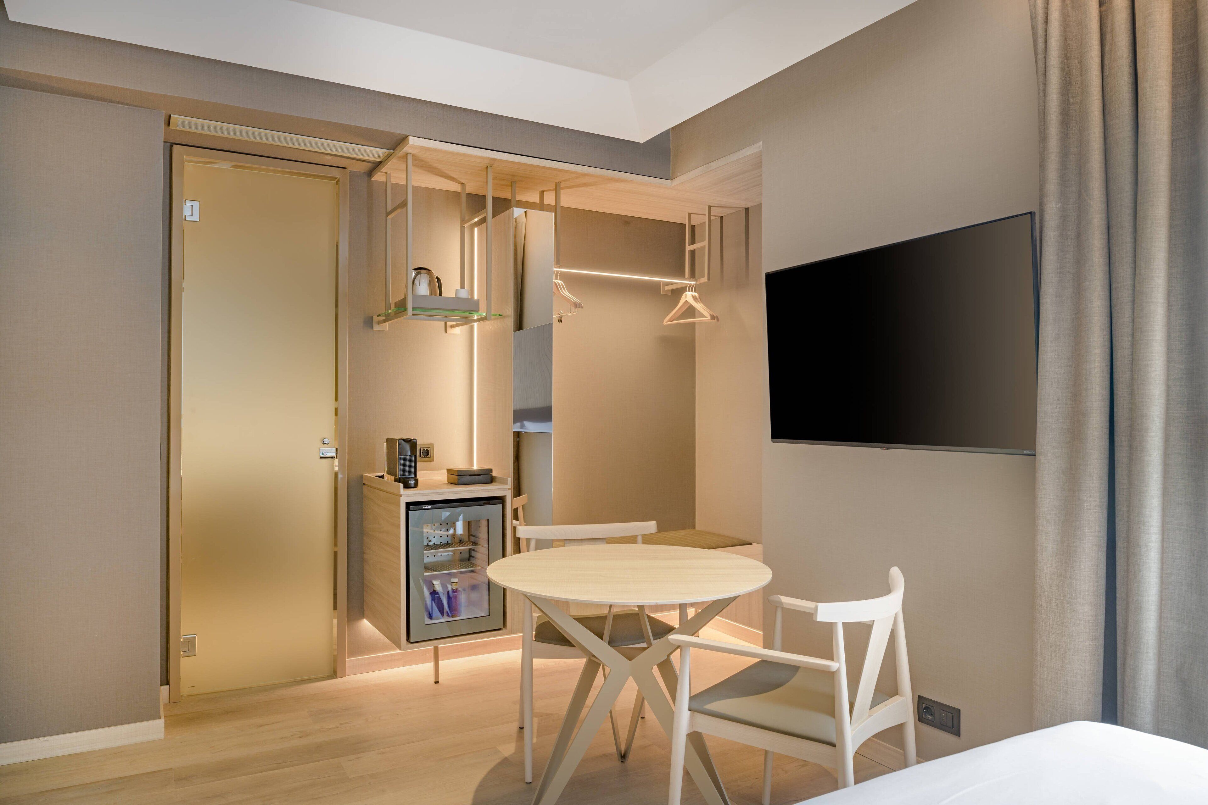 Una habitación de hotel moderna con una mesa de comedor de madera, sillas blancas, un televisor de pantalla plana y un mini bar.