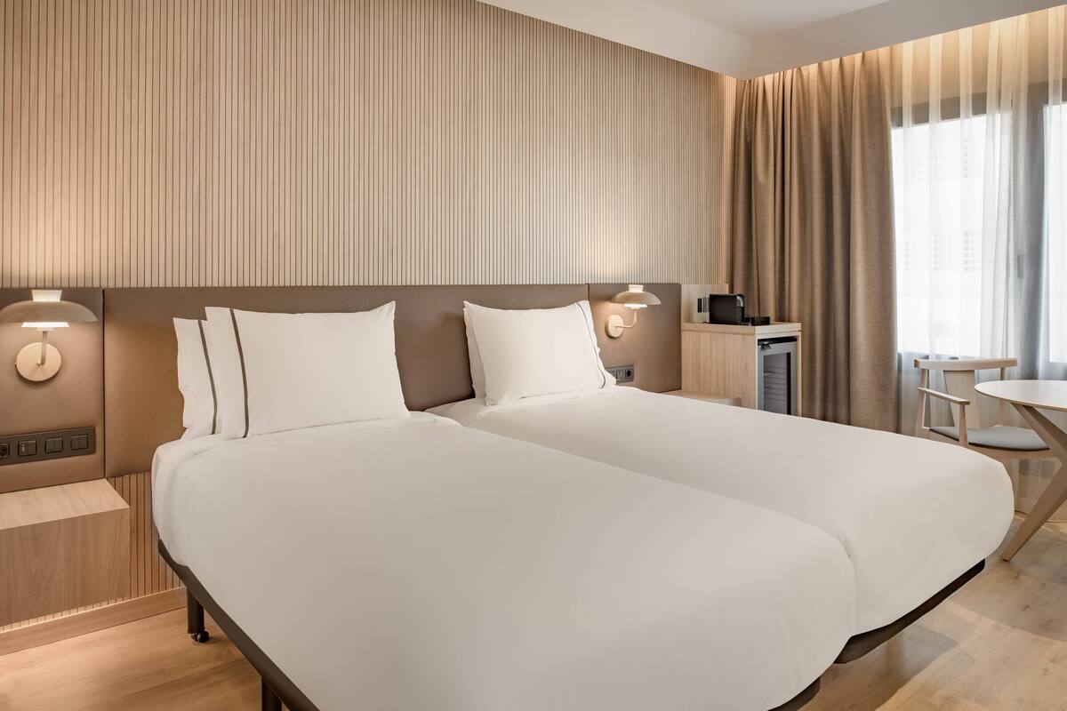 Ein Hotelzimmer mit einem großen Bett, einem Nachttisch, einer Lampe und Blick auf die Stadt durch das Fenster.