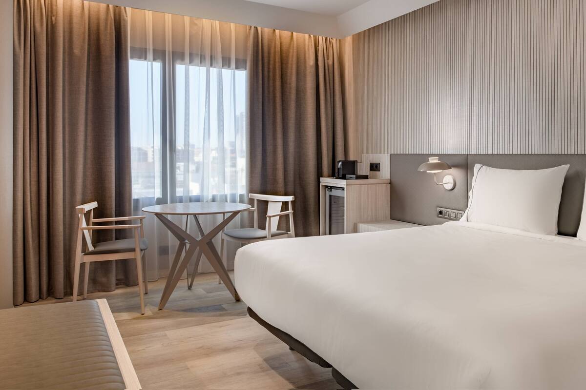 Ein Hotelzimmer mit einem großen Bett, einem Schreibtisch mit Stuhl, einem kleinen Tisch und Blick auf die Stadt durch große Fenster.