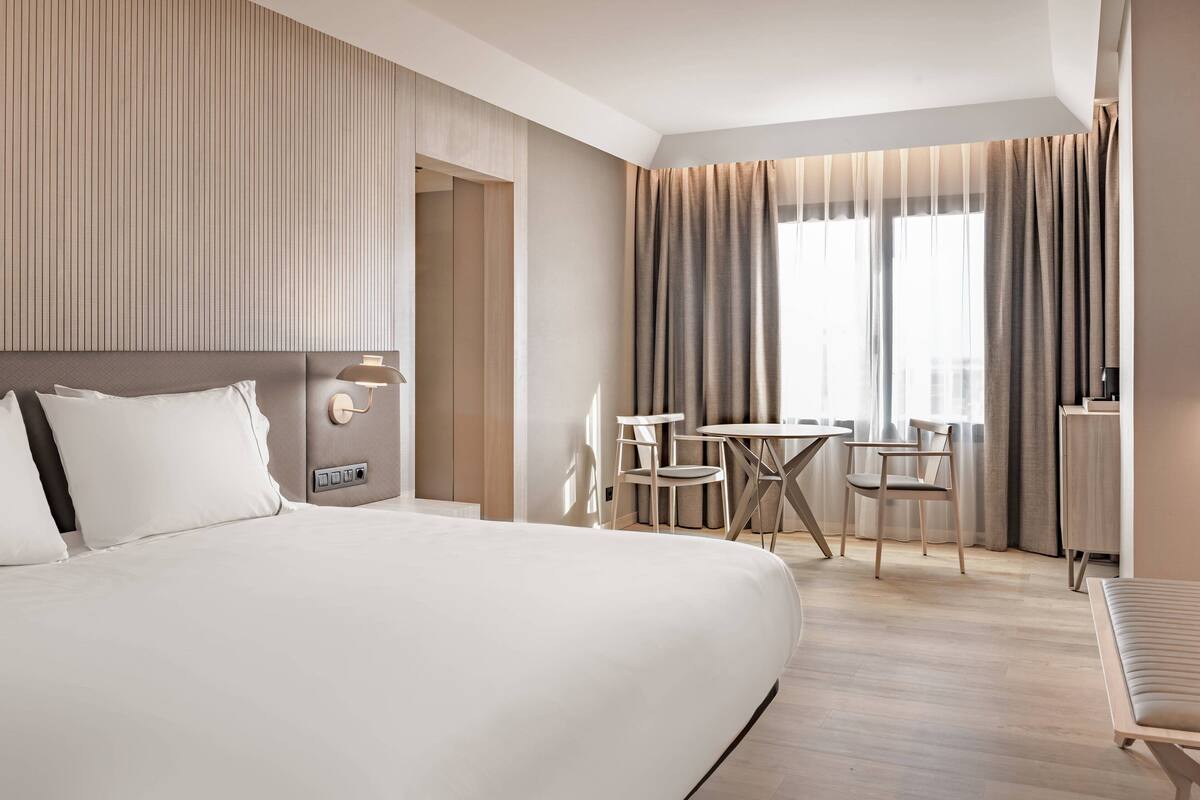 Une chambre d’hôtel moderne dotée d’un grand lit, d’un bureau et de chaises, d’une petite table et offrant une vue sur la ville grâce à de grandes fenêtres.