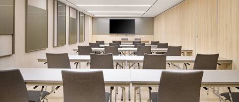 A conference room with a long table, multiple chairs, a flat-screen TV, and wooden panel walls.