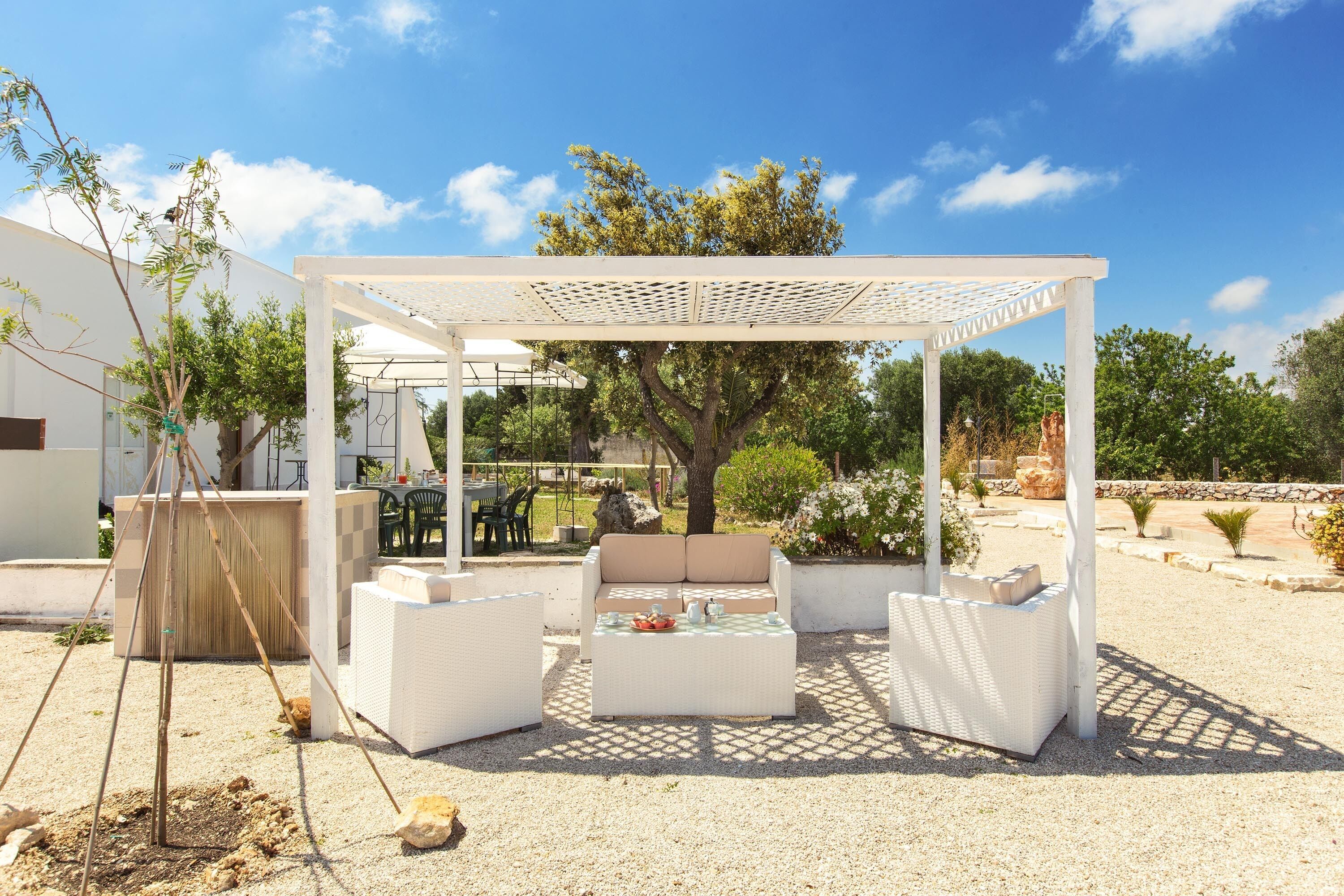An outdoor seating area with white wicker furniture, a pergola, and a tree in the background.