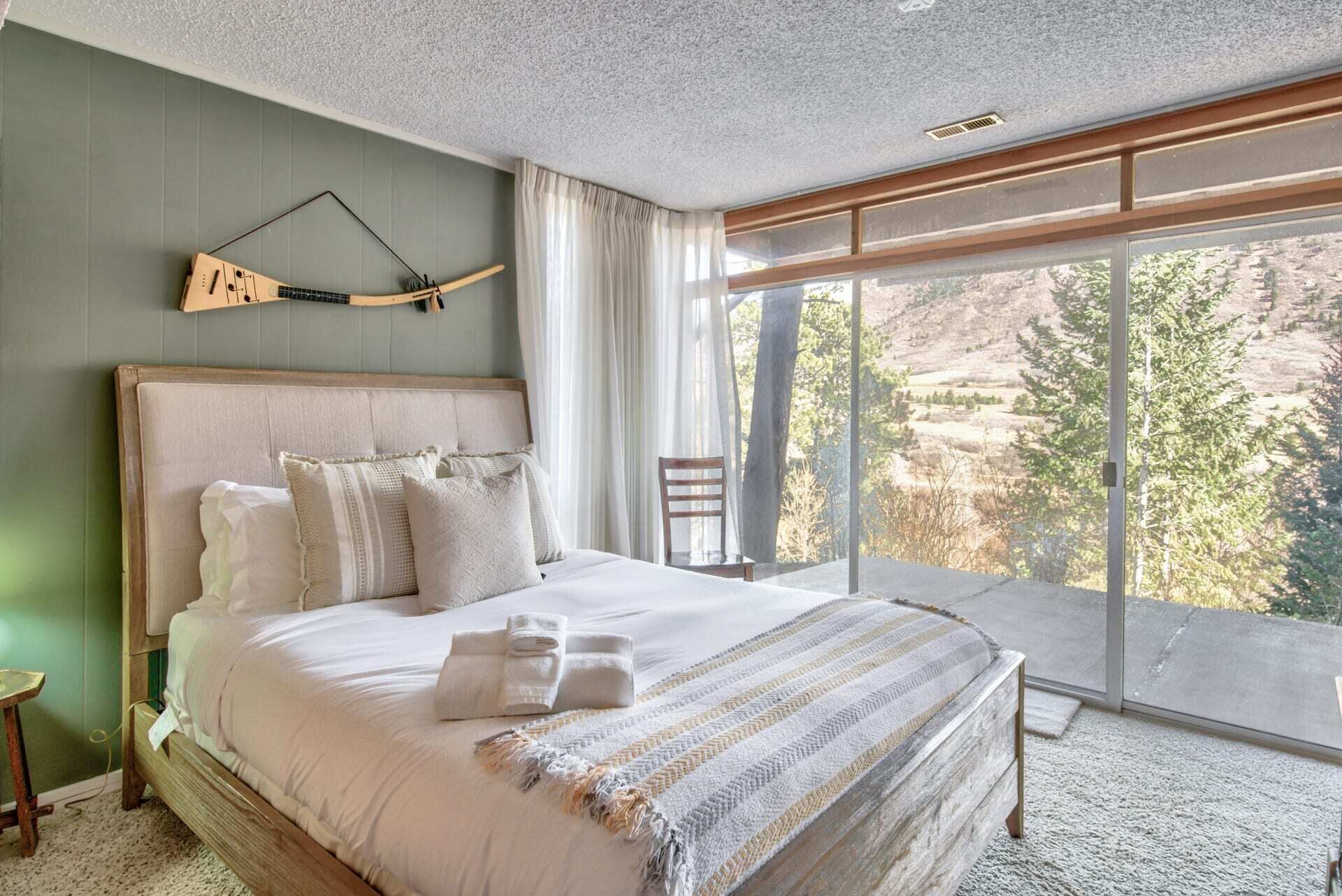 Un dormitorio con una cama grande, un cabecero de madera, vistas al exterior a través de amplias ventanas y una guitarra colgada en la pared.
