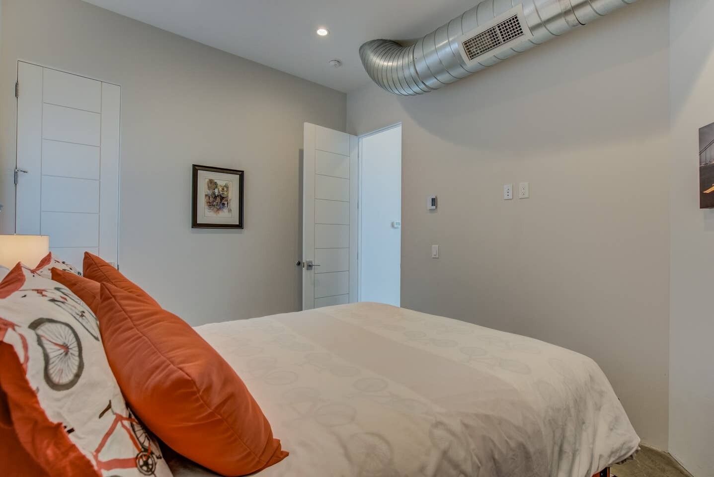 Une chambre à coucher avec un lit, deux portes blanches, un tableau encadré accroché au mur et une bouche d’aération fixée au mur.