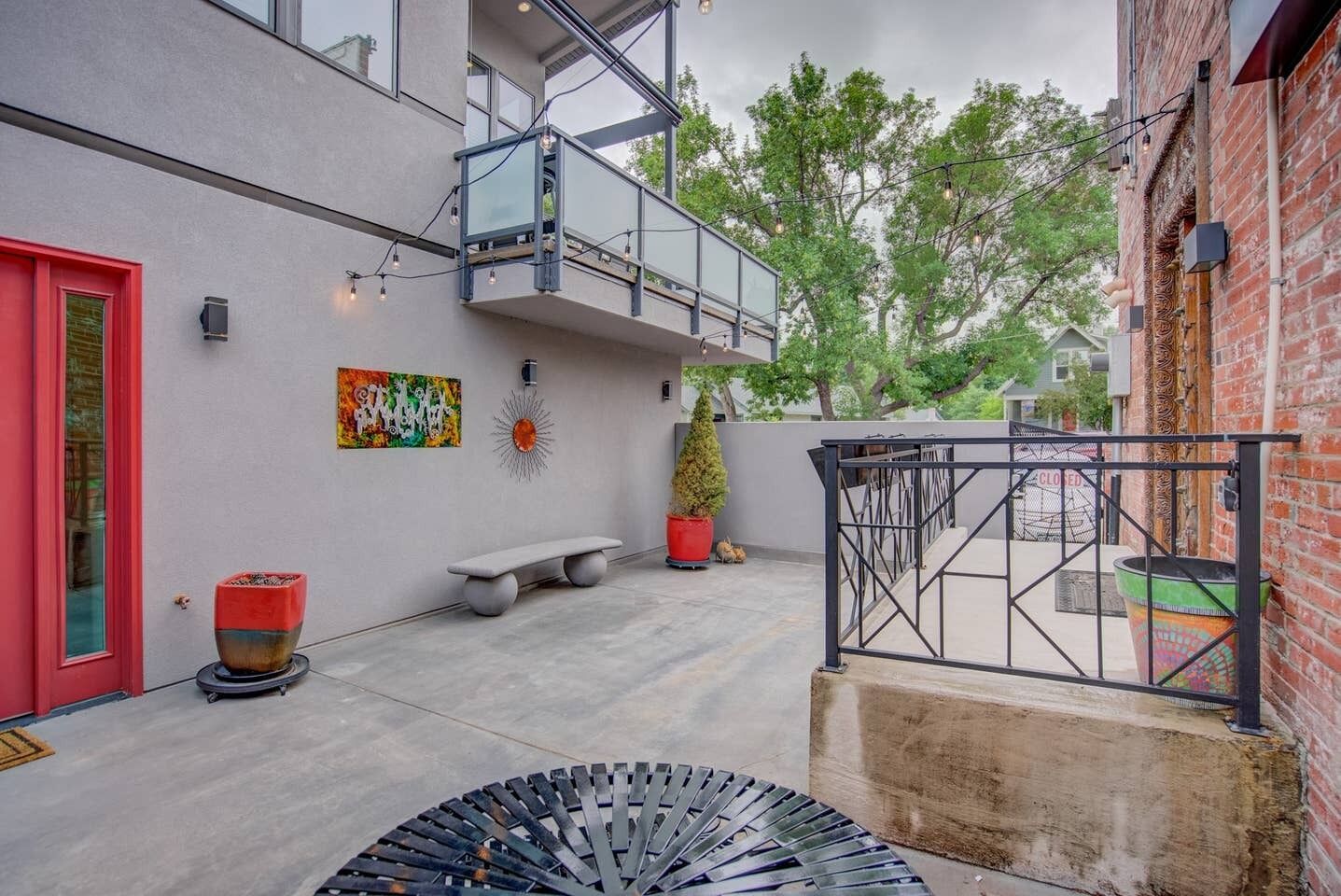 An outdoor courtyard with a red door, a bench, a potted plant, a metal sculpture, and a closed gate.