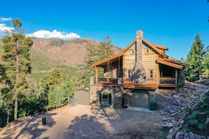 A log cabin with a stone chimney, a porch, and a view of mountains.