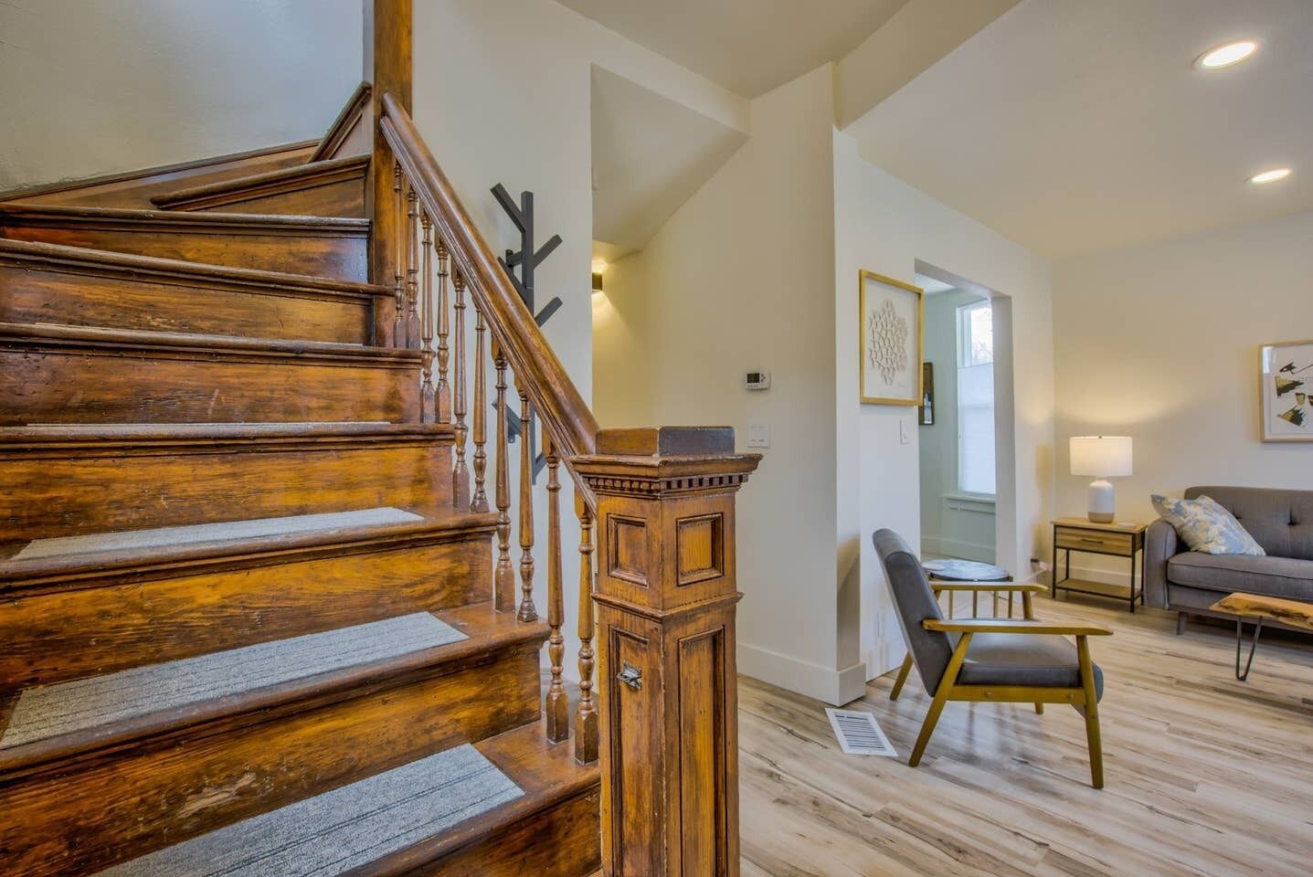Eine Holztreppe mit Handlauf, ein Holzkasten, ein graues Sofa, ein kleiner Tisch mit Lampe und ein Sessel in einem Wohnzimmer mit Holzdielen.