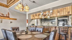 Un coin repas avec une table en bois et des chaises en cuir, une cuisine avec des meubles en bois et une hotte de cuisinière en acier inoxydable, et un plafond avec un revêtement texturé et des luminaires suspendus.
