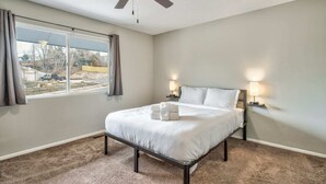 Un dormitorio con cama, ventilador de techo, dos lámparas de noche, ventana con cortinas y vistas al exterior.