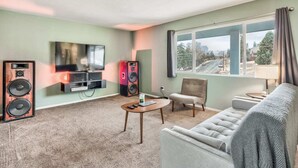 Una sala de estar con televisor, parlantes, un sofá, una silla y una ventana con vista a la calle.