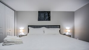 Cama bem arrumada com lençóis brancos, duas luminárias de cabeceira e janela com cortina escura.