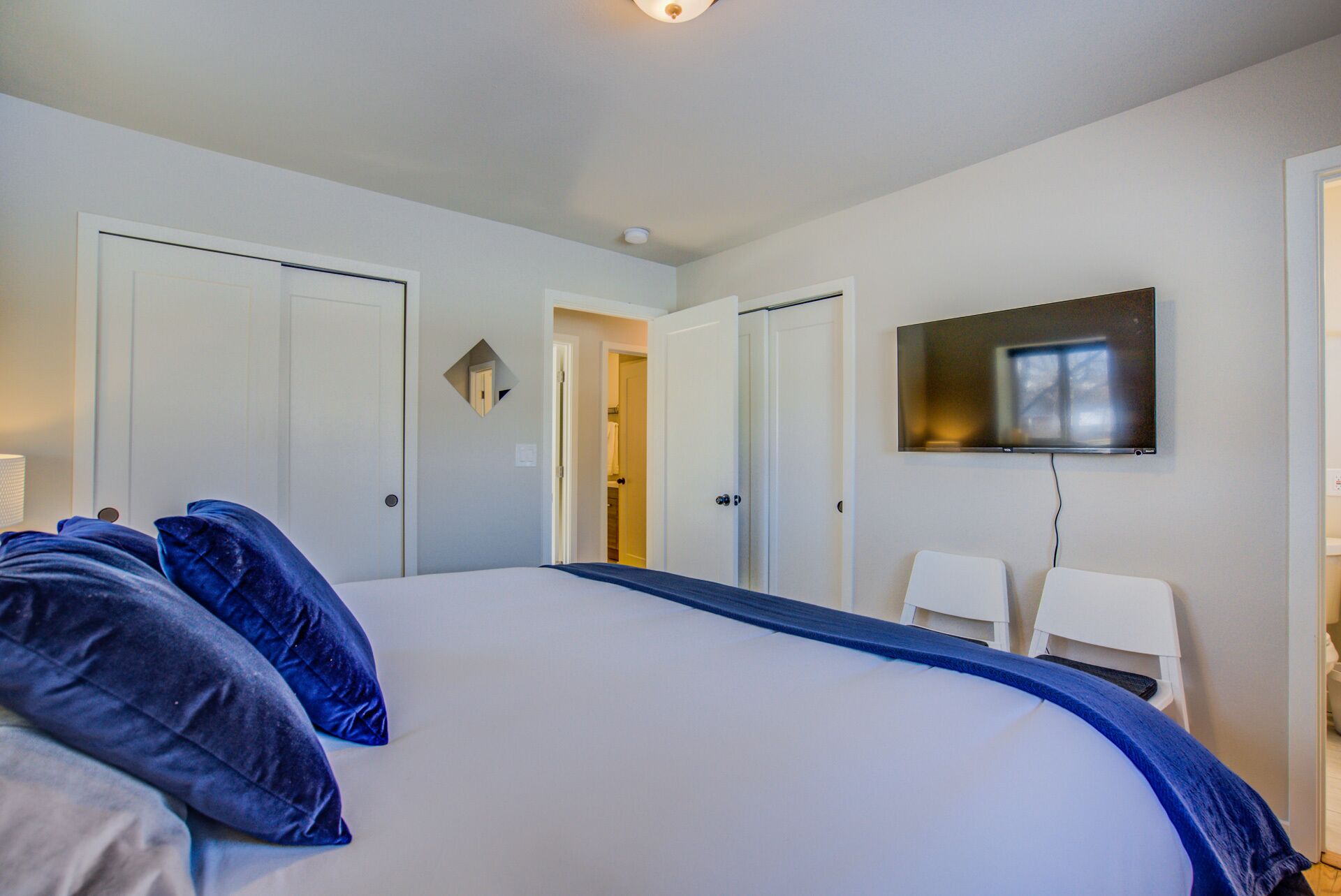 A bedroom with a large bed, white walls, a TV mounted on the wall, and a door leading to another room.