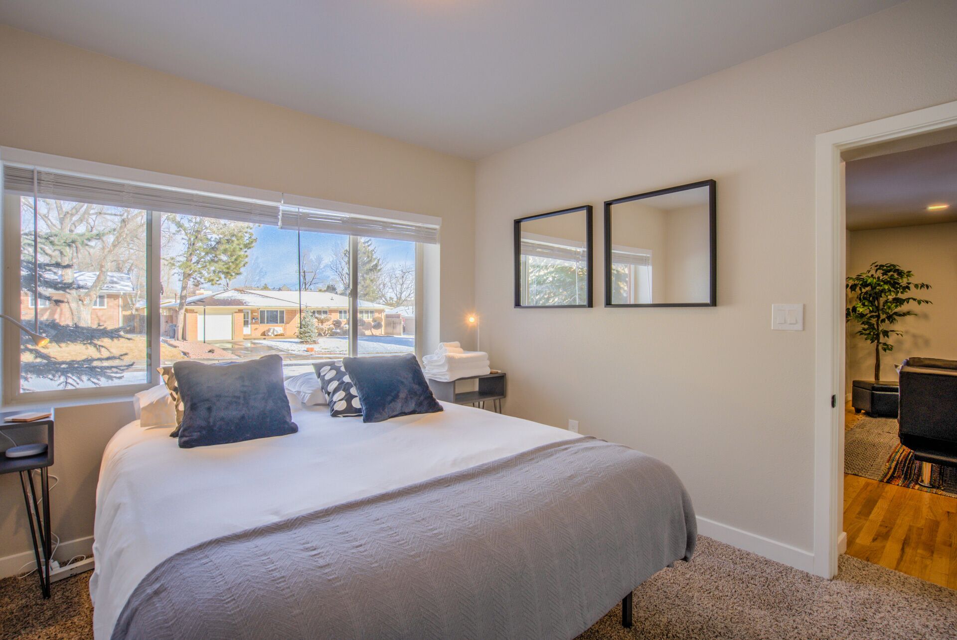 A bedroom with a large bed, a window with a view of a house, a desk with a chair, and a mirror on the wall.