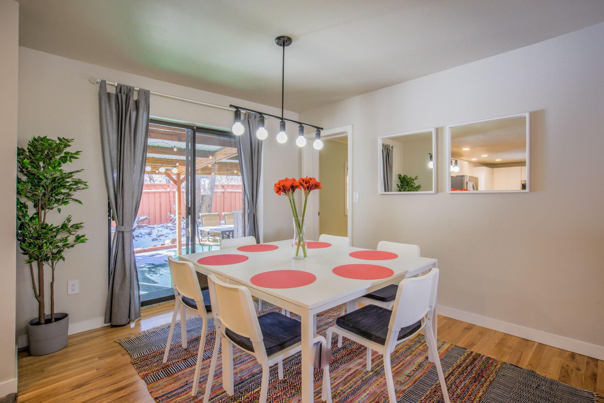 Un comedor con una mesa blanca, individuales circulares rojos, sillas blancas, un jarrón con flores rojas y una lámpara colgante.