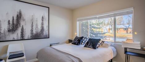 A bedroom with a bed, a chair, a desk, a window with a view of the outside, and a painting on the wall.
