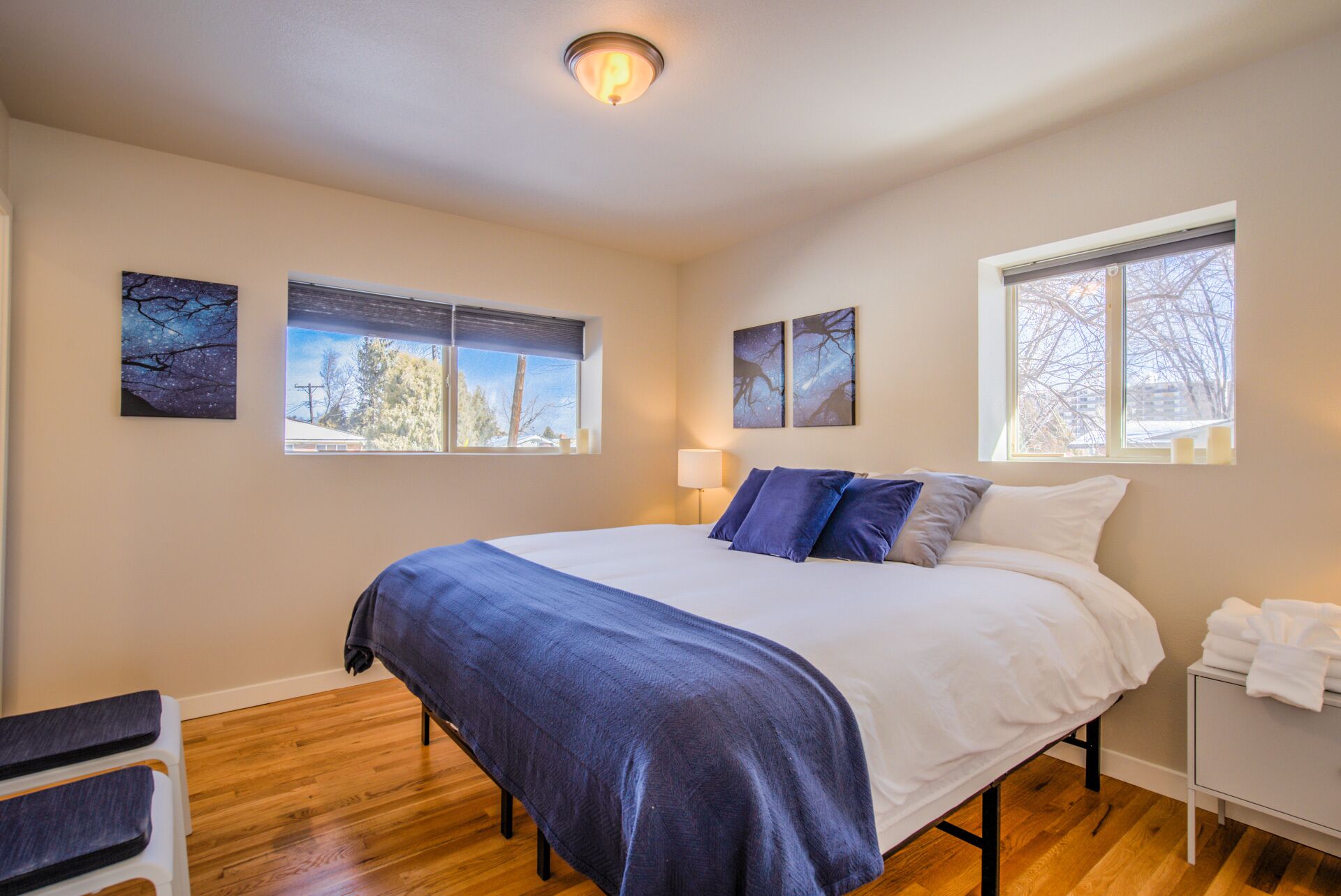 A bedroom with a large bed, two windows, a bedside table with a lamp, and artwork on the walls.