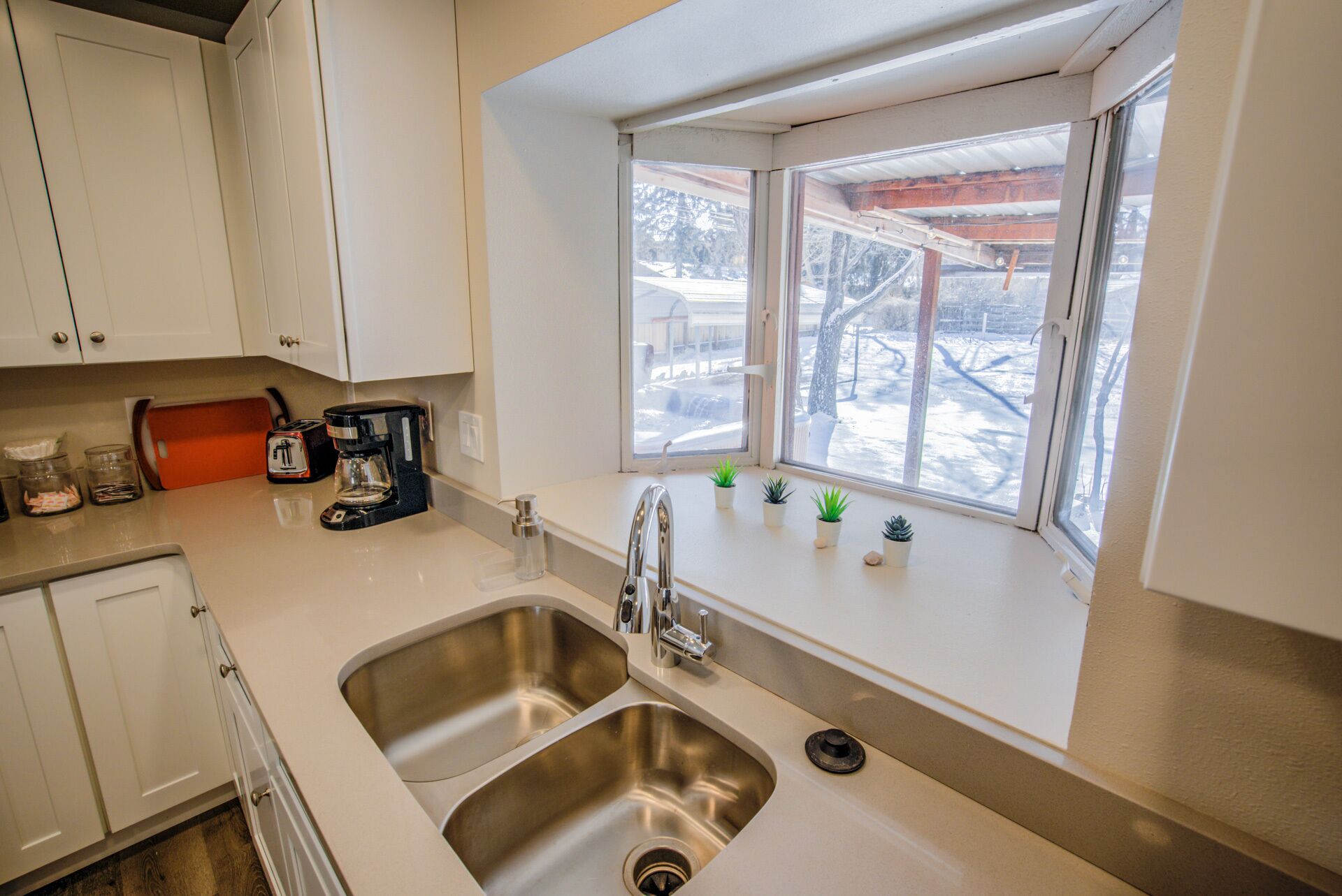 A kitchen with a double sink, white cabinets, a coffee maker, and a window with a view of a snowy outdoor area.