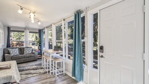 A cozy living room with a sofa, a coffee table, and a dining area with a table and chairs. There are large windows with curtains, a door, and a ceiling light fixture.