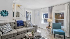 A living room with a grey sofa, a blue armchair, a small table with a lamp, and a large window with curtains.