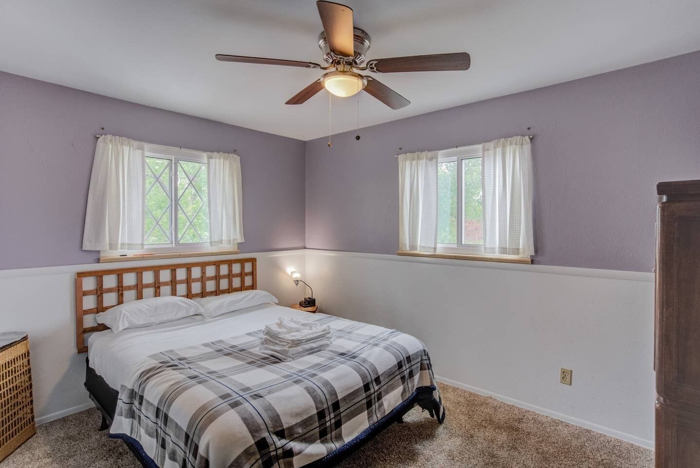 Quarto com cama, duas janelas com cortinas, ventilador de teto e mesa de cabeceira com abajur.
