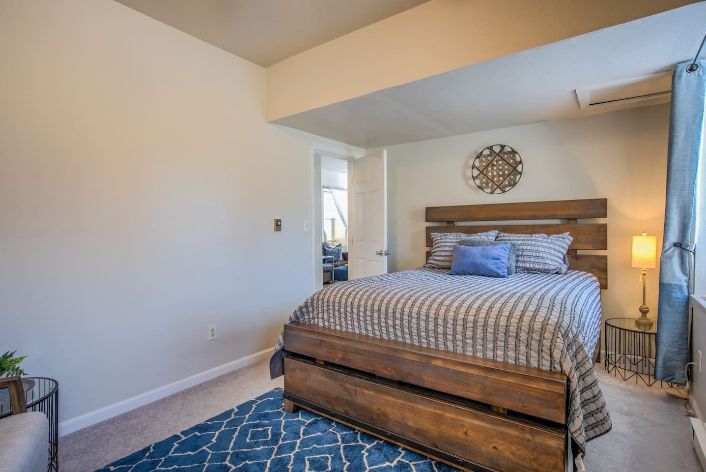 A bedroom with a wooden bed, a blue patterned rug, a small bedside table with a lamp, and a decorative wall piece.