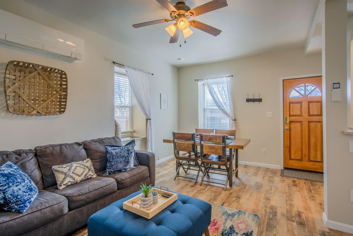 A living room with a brown leather sofa, blue ottoman, and a wooden dining table with chairs.