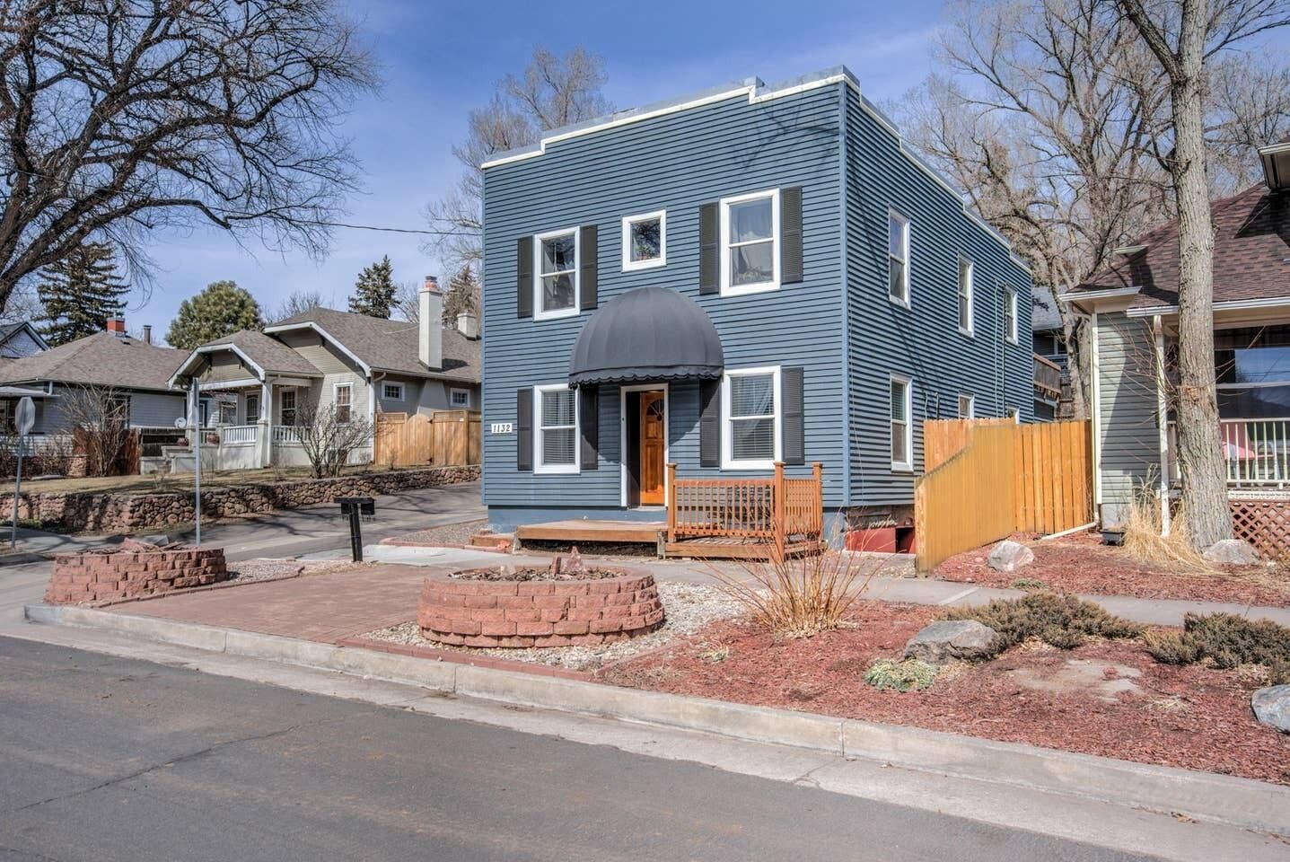 A two-story house with a front porch, a brick fire pit, and a wooden fence.
