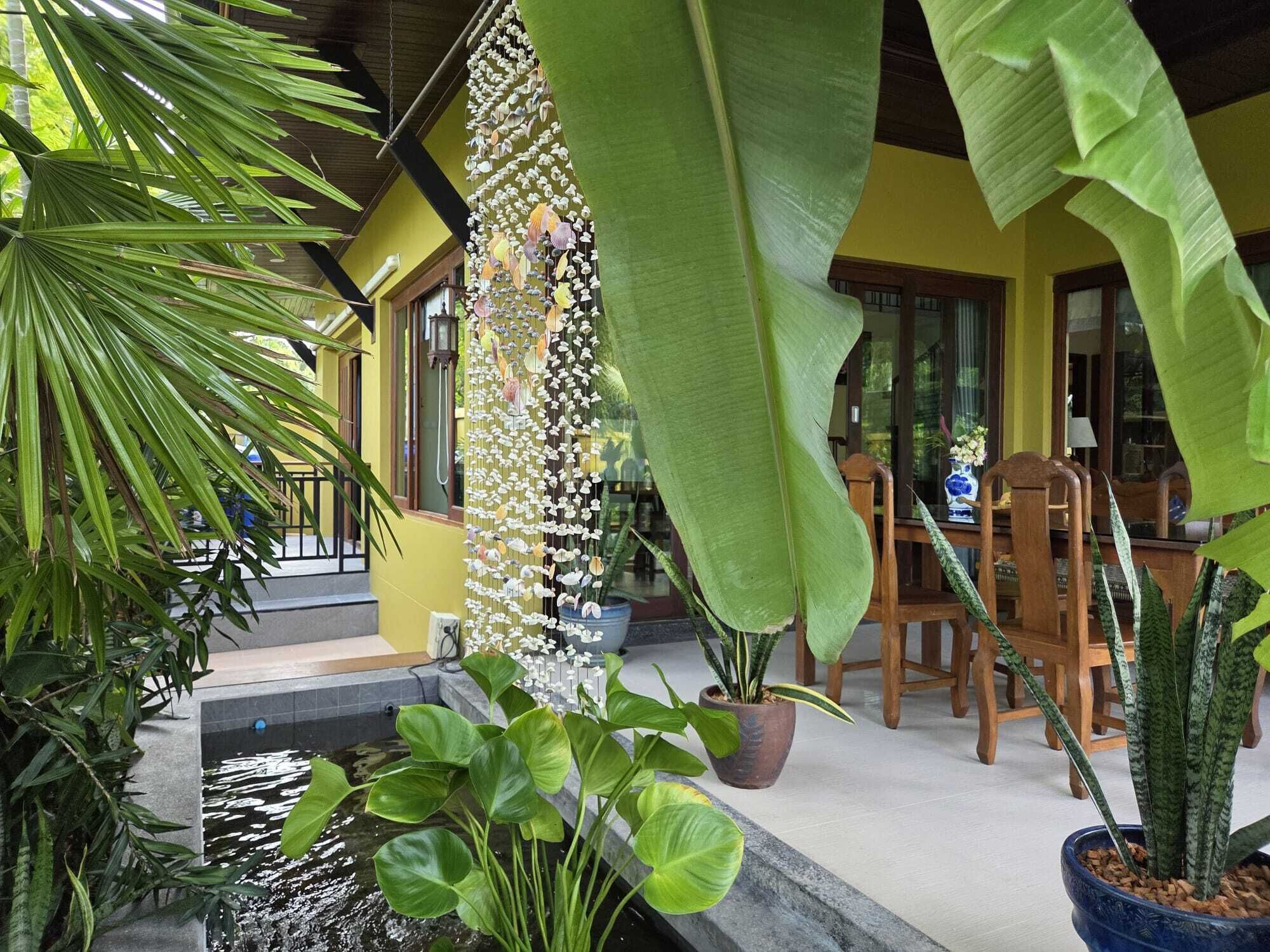 A tropical garden with a water feature, potted plants, and a wooden table and chairs.